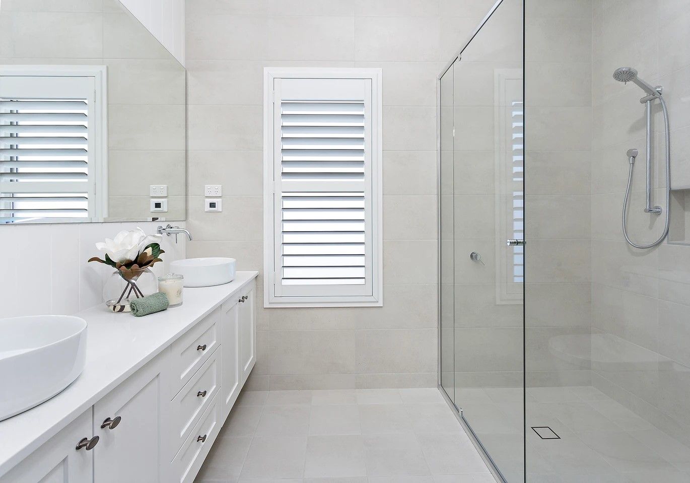 White bathroom with a long vanity, sinks, shower, and shuttered windows.