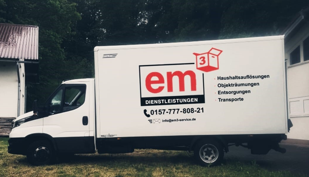 Ein weißer Lastwagen mit dem Wort „em3“ auf der Seite steht vor einem Gebäude.