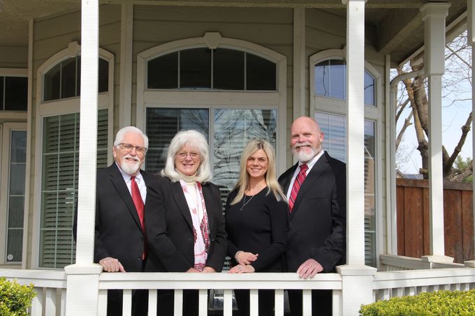 Four people in formal attire stand on a porch in front of a house, smiling at the camera.