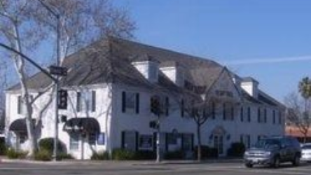 White multi-story building with dark roof and dormers on a street corner, with parked cars and trees nearby.
