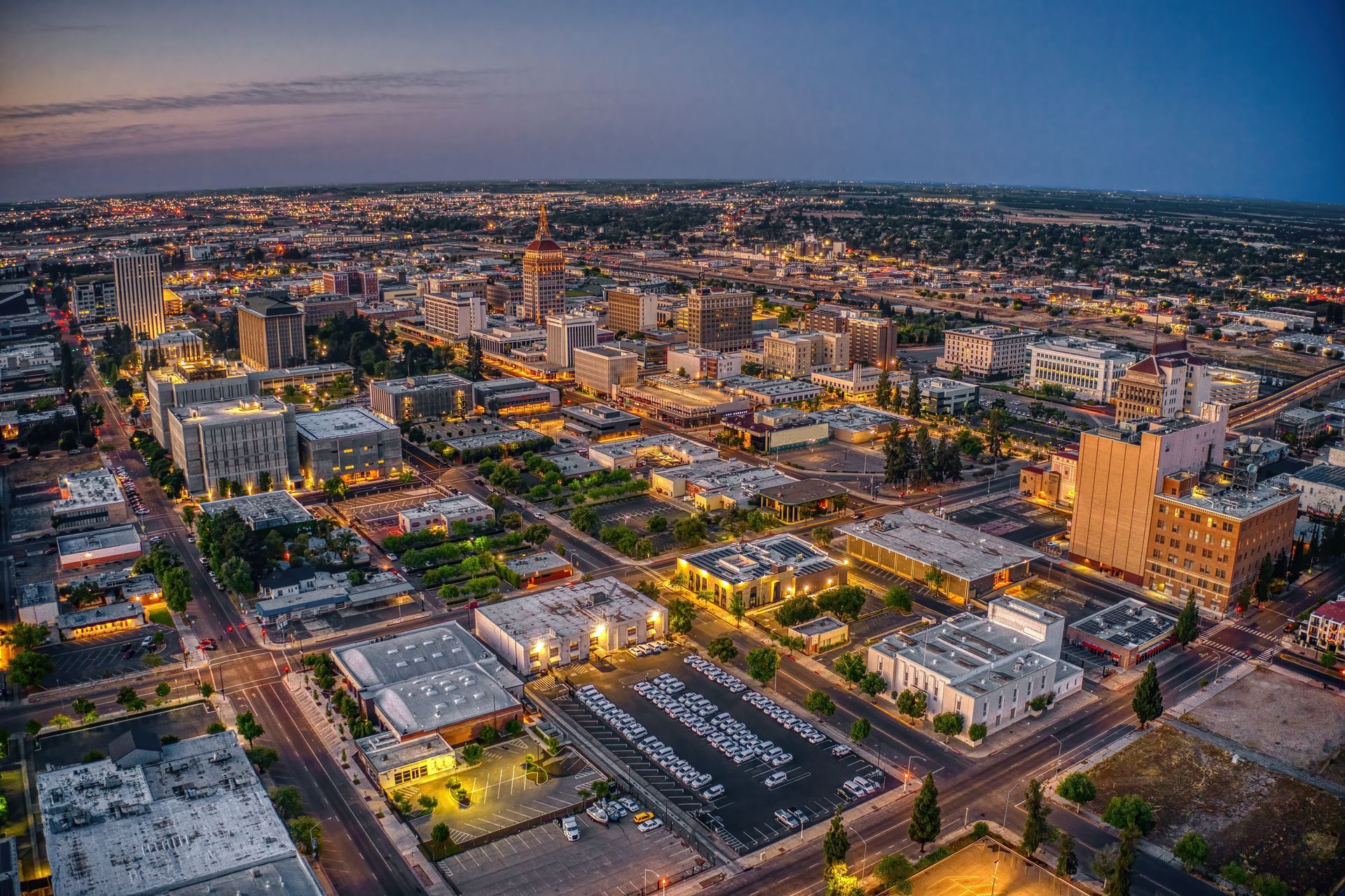 Aerial view of a city at dusk with glowing streets, buildings, and a pink-blue sky.