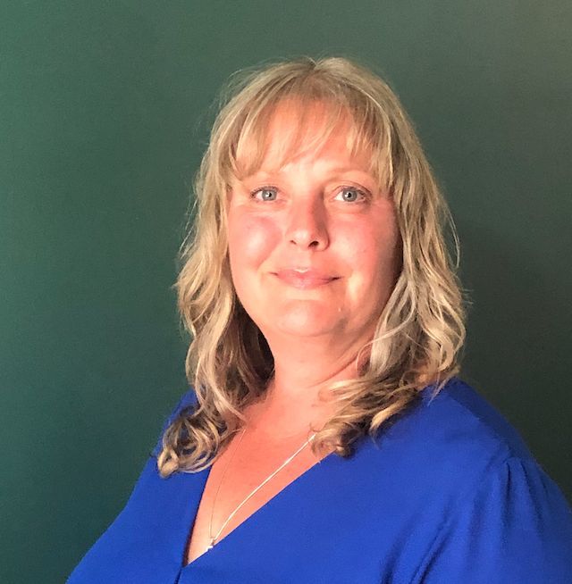 Blonde woman in blue top smiles at camera in front of a green background.