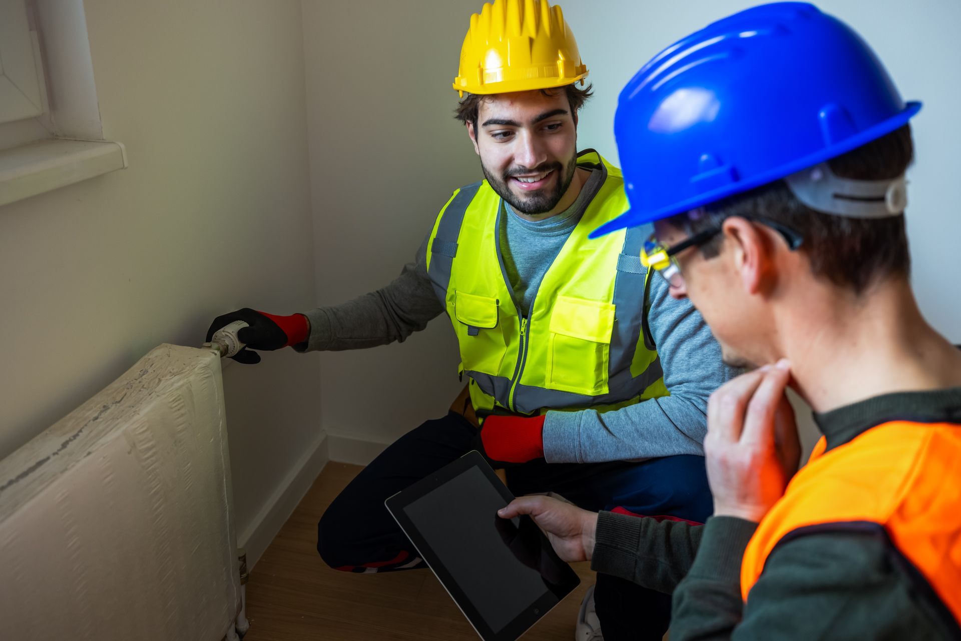 Two heating contractors in hard hats and safety vests inspecting a home heater with a tablet Two heating contractors in hard hats and safety vests inspecting a home heater with a tablet