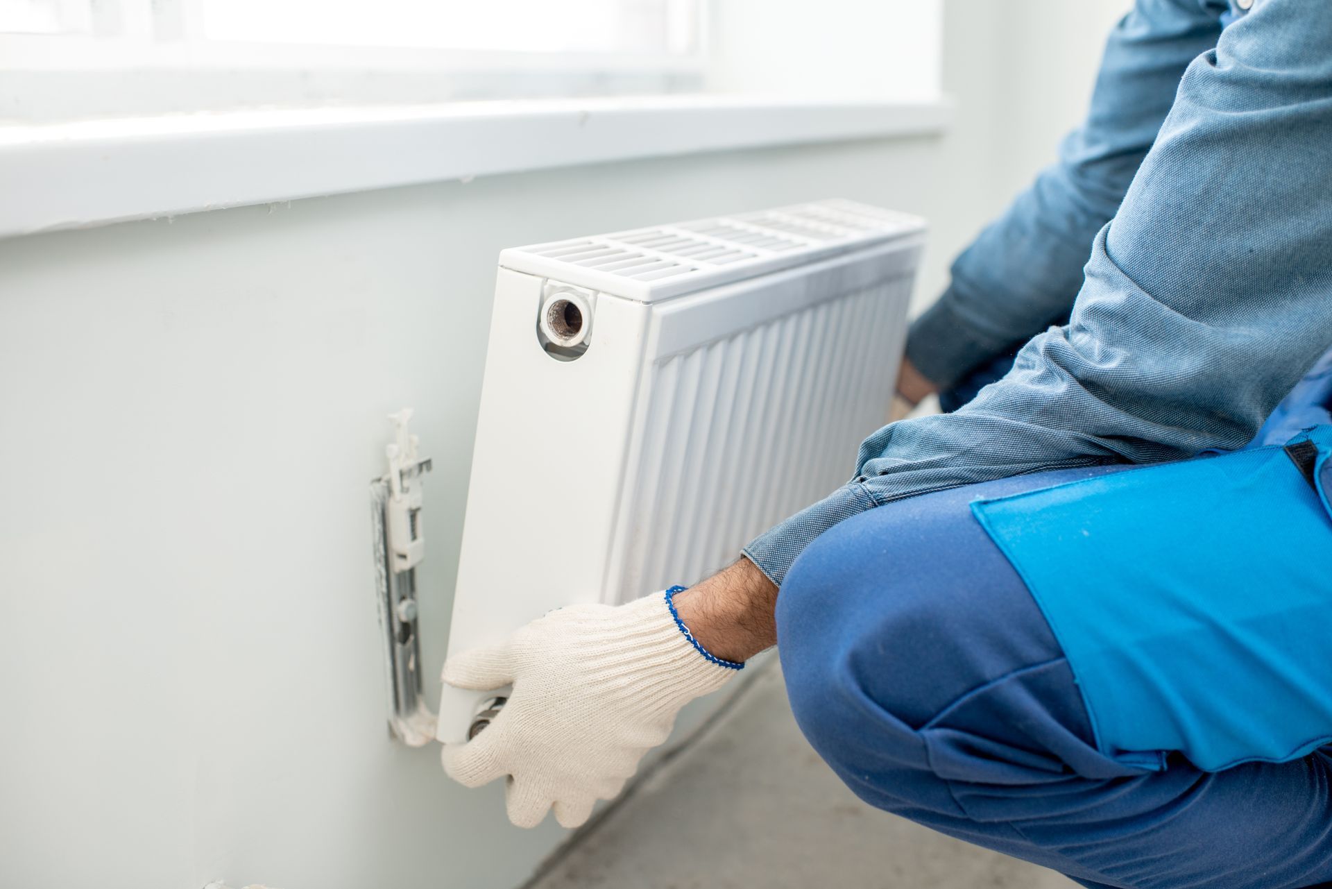 A technician in a blue uniform installing a white wall-mounted heating radiator under a window