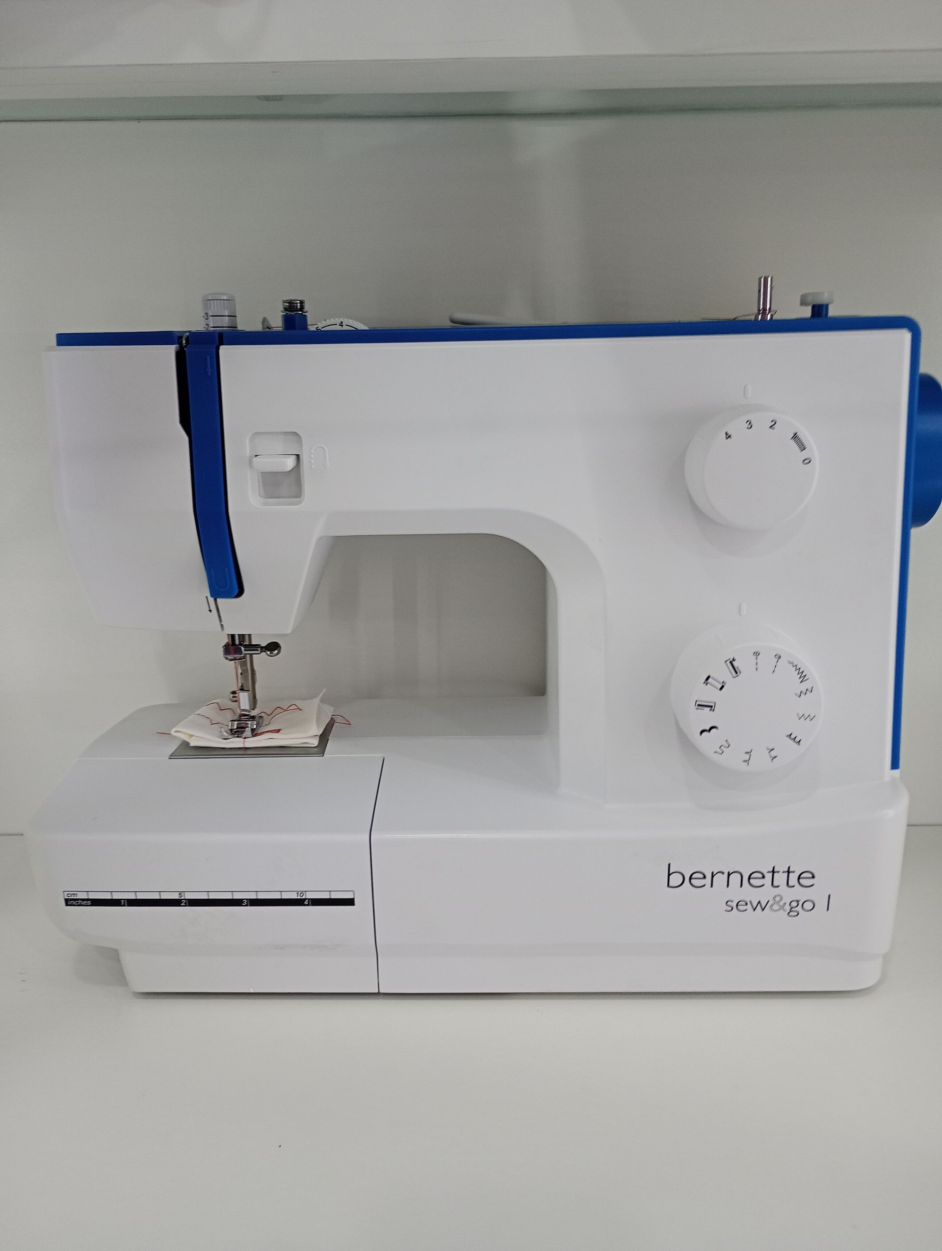 Máquina de coser blanca y azul en un estante. La máquina está cosiendo un pequeño cuadrado de tela.
