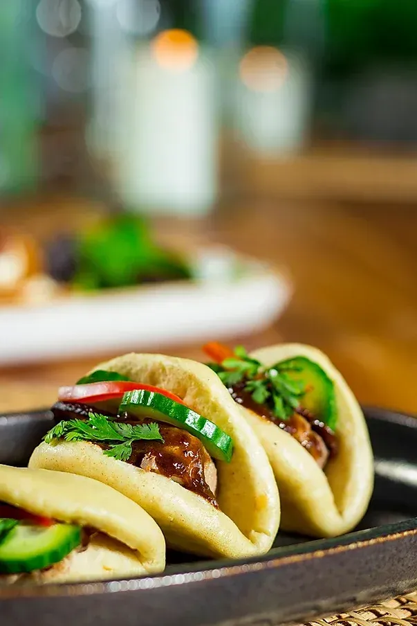 Three bao buns filled with meat, cucumber, and cilantro, on a dark plate.