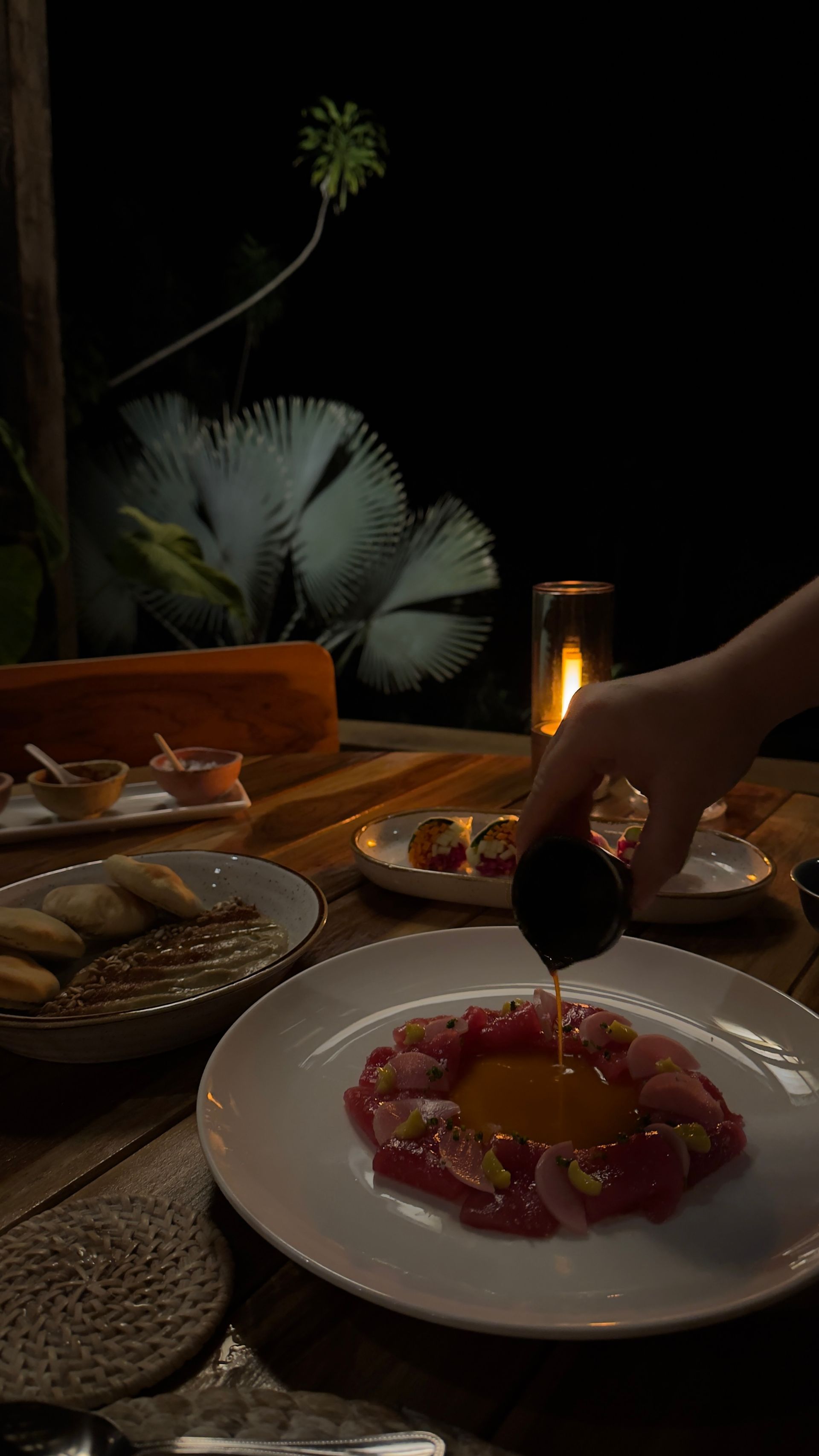 A hand pouring sauce over a dish of raw fish, outdoors at night. Candlelit table, other food nearby.