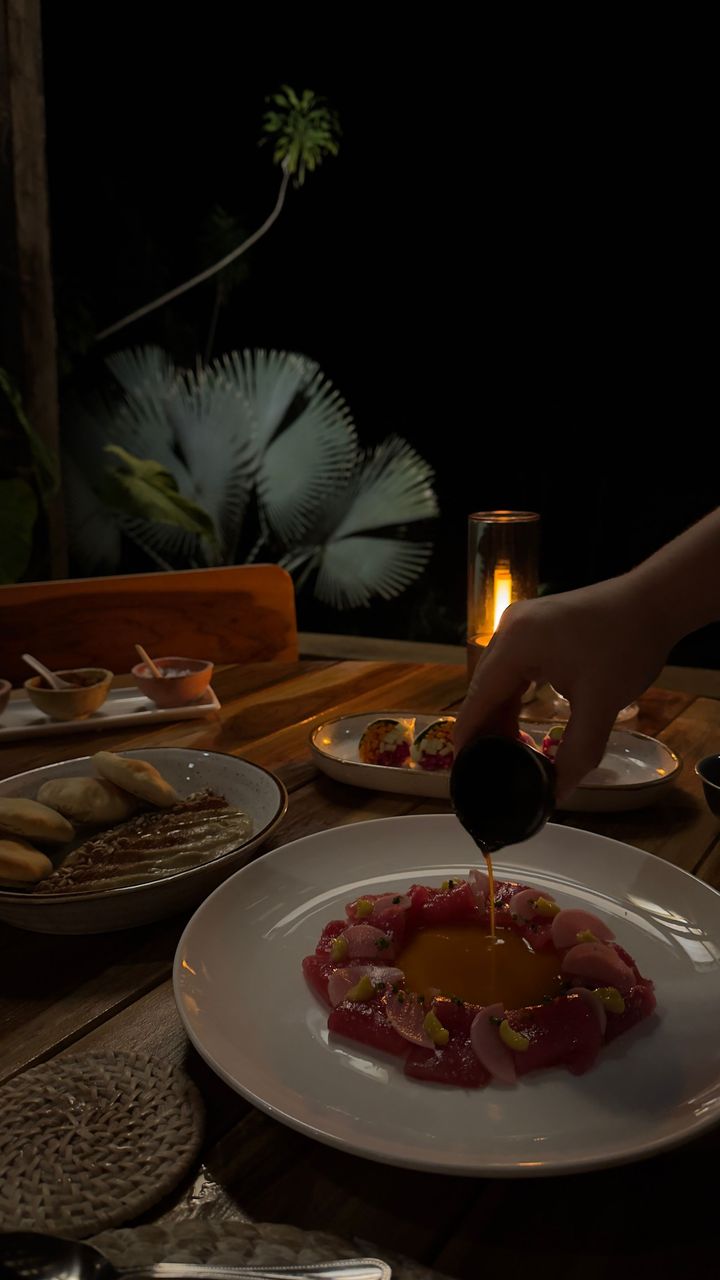 A hand pouring sauce over a dish of raw fish, outdoors at night. Candlelit table, other food nearby.