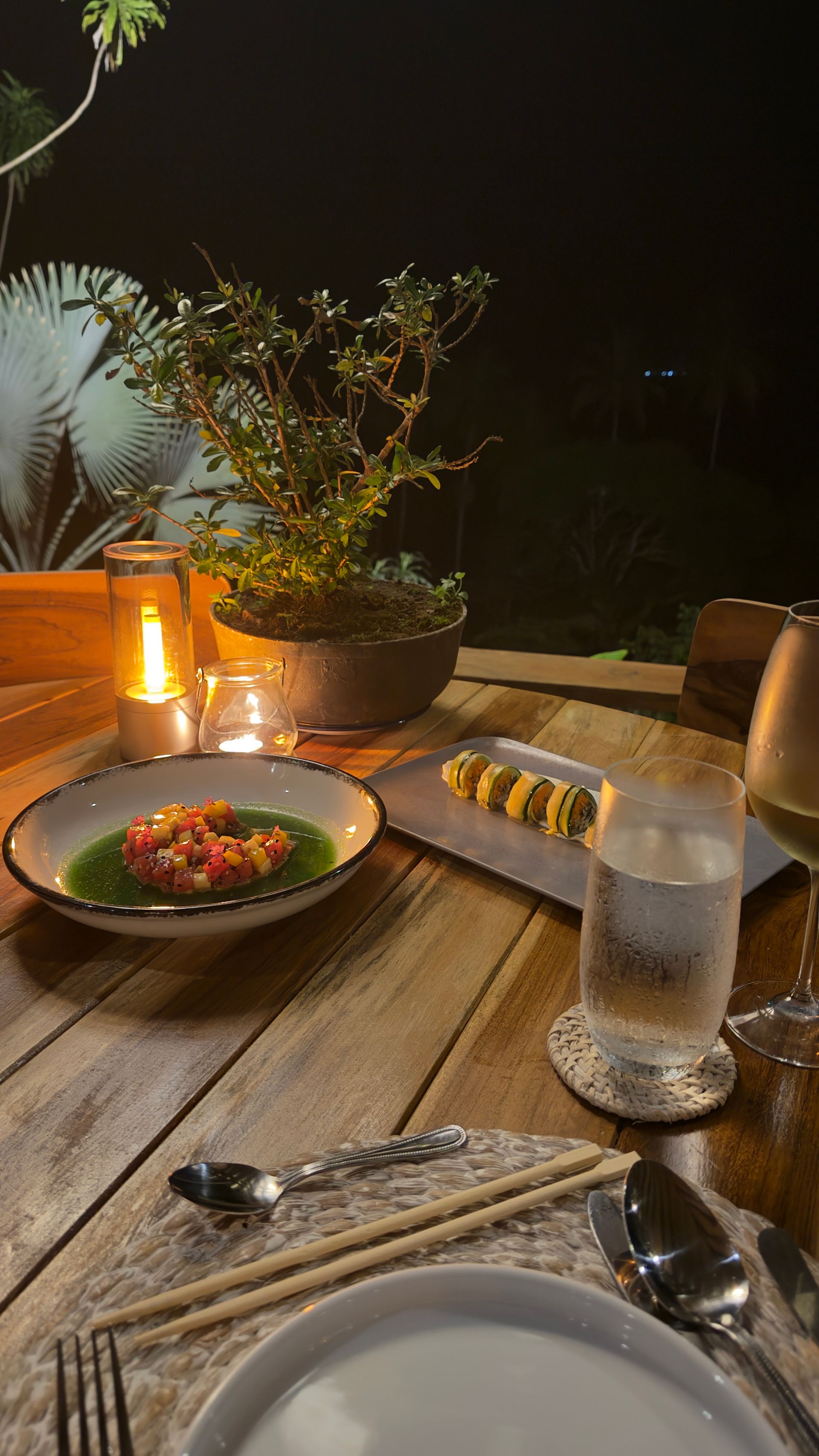 Dinner table set outdoors at night with food, candles, and plants.
