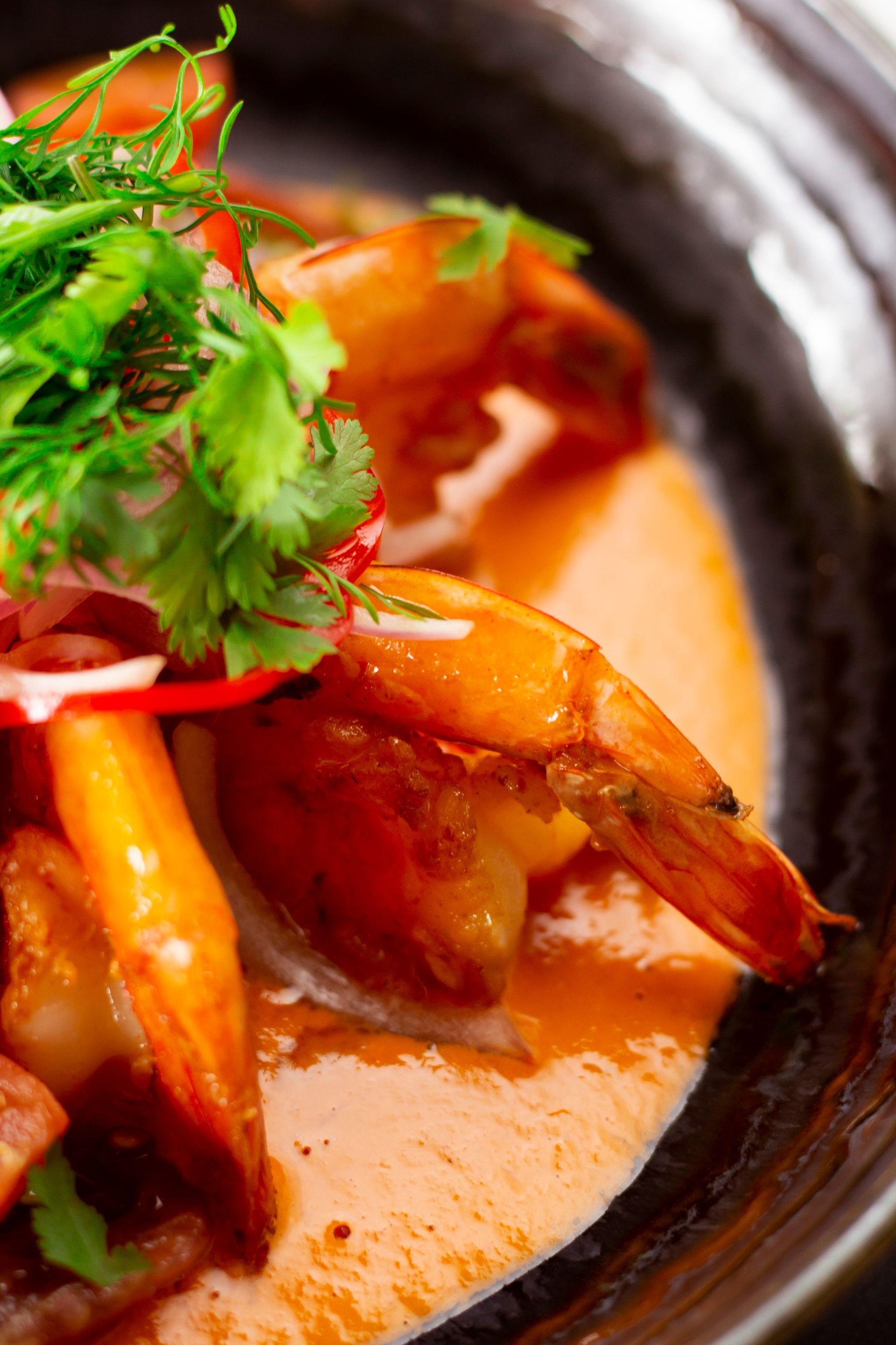 Shrimp in a sauce, garnished with cilantro and radish, in a dark bowl.
