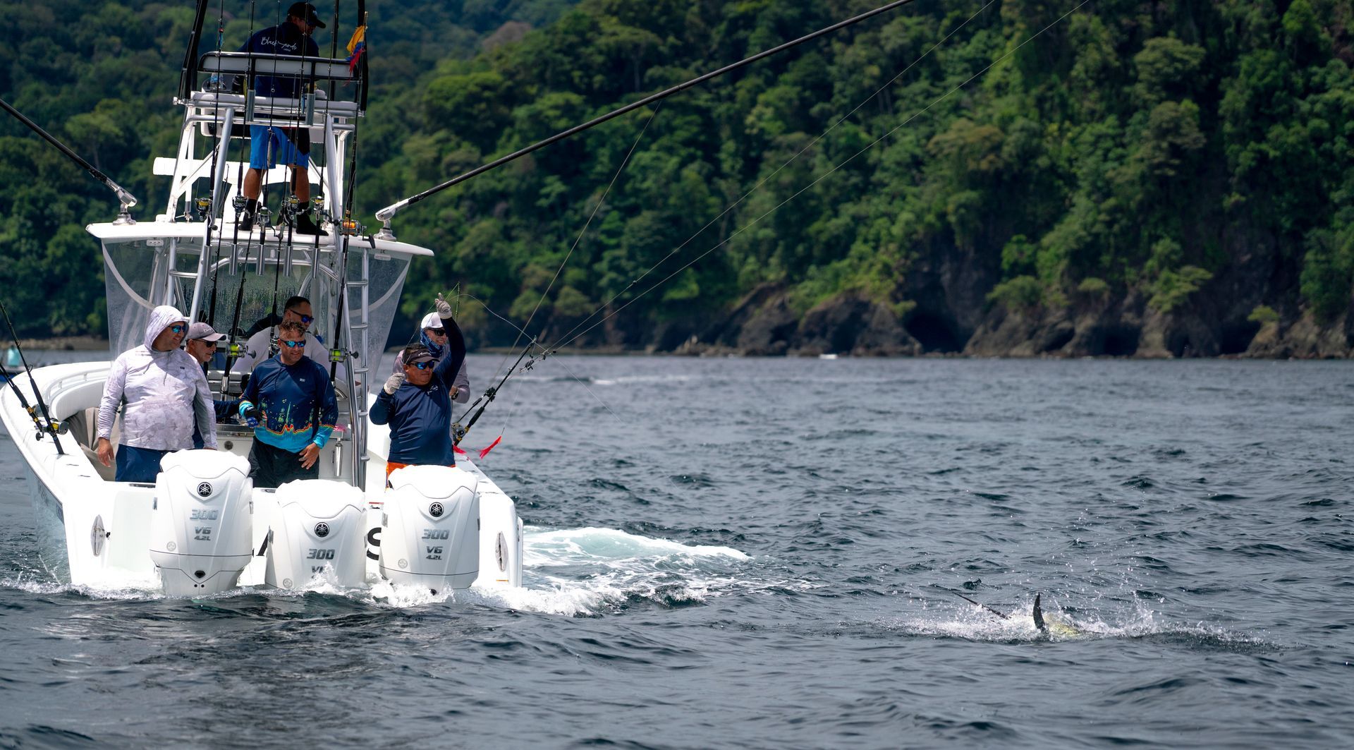 Fishing boat with people reeling in a fish in blue water near a forested shore.