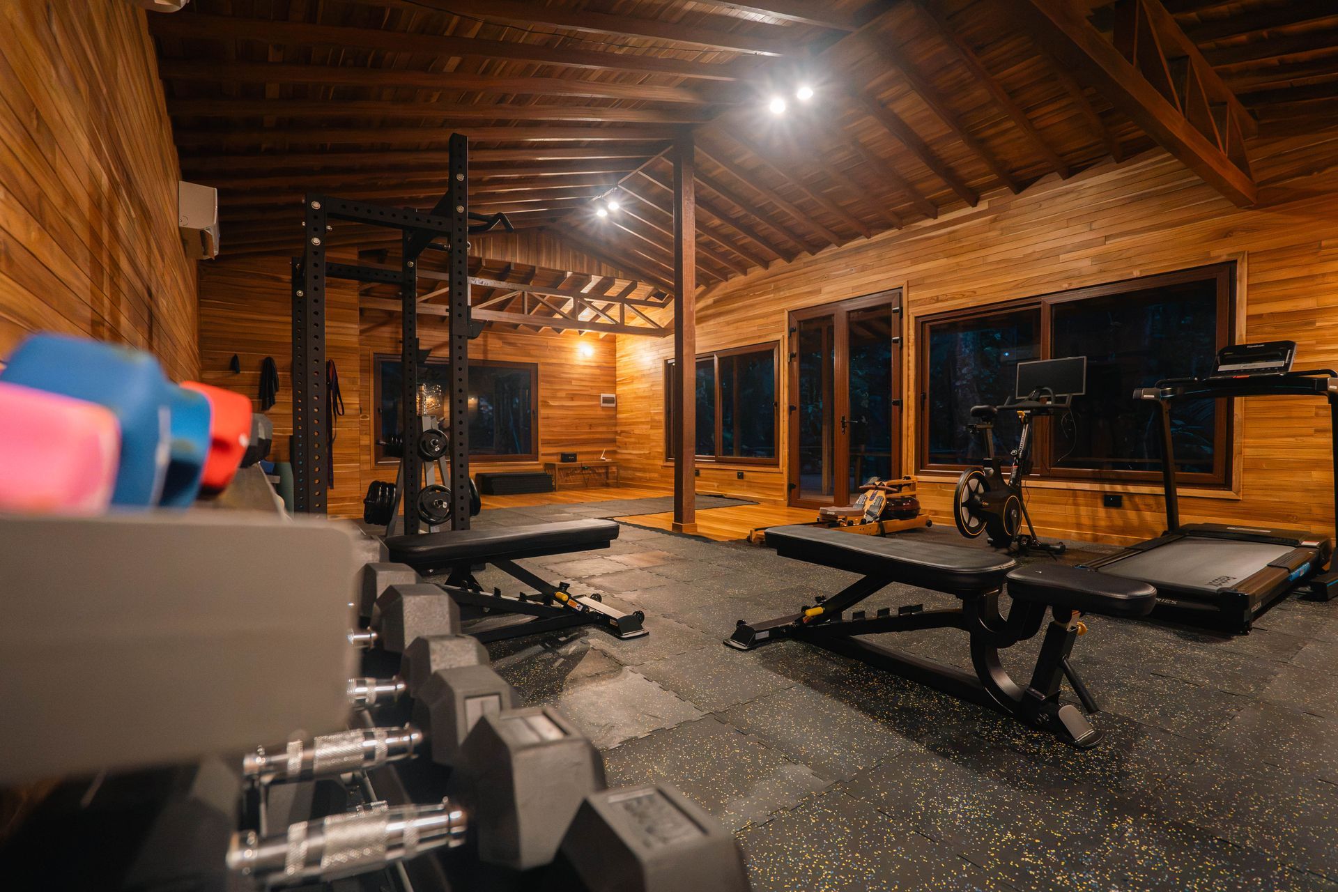 Wooden-walled home gym with various exercise machines, weights, and a rowing machine. Window view of trees.