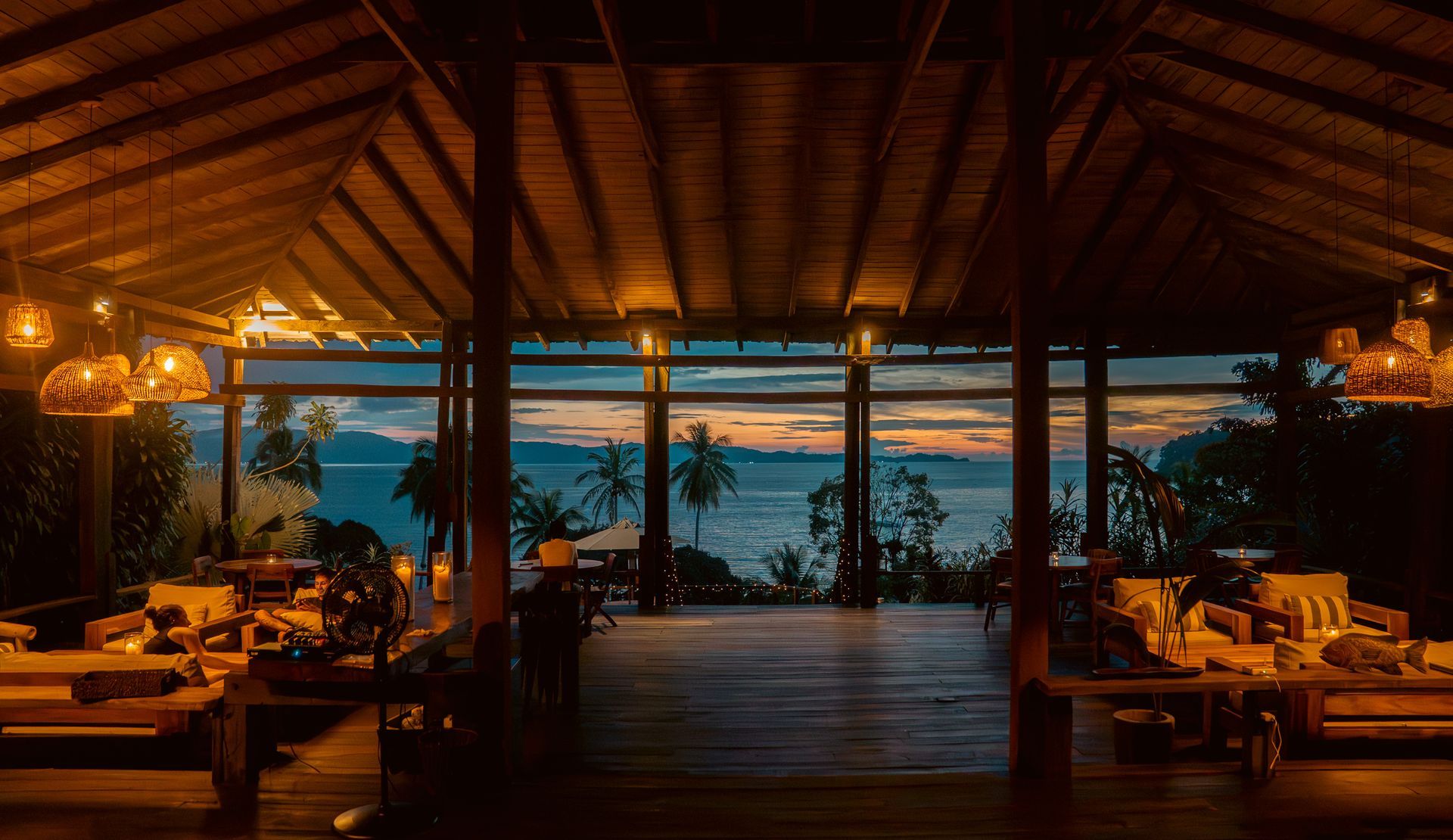 Covered outdoor dining area, wooden furniture, hanging lights, lush green foliage in background.