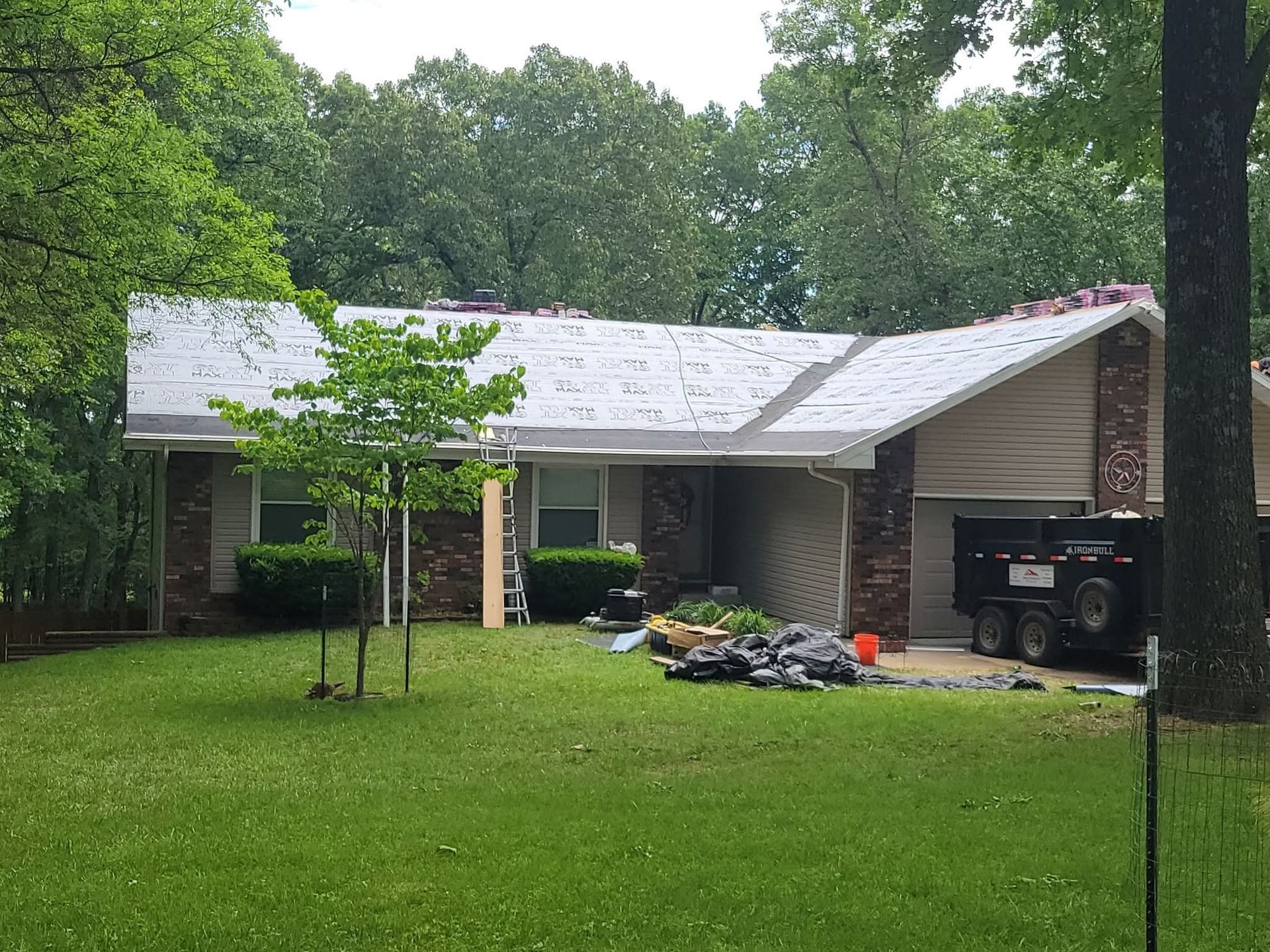 House with partially replaced roof. White tarp covers, bricks, siding, surrounded by trees.