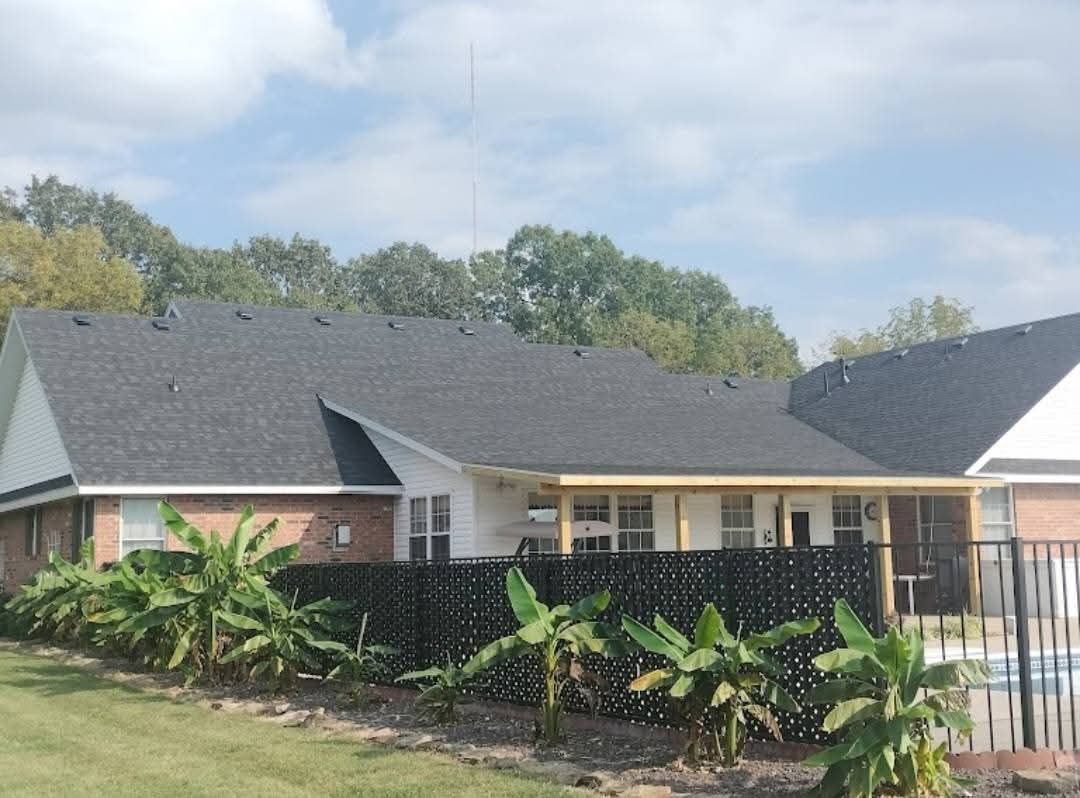 A home with a dark roof and a fence made of bottles, in front of banana plants. Trees and a blue sky are in the background.