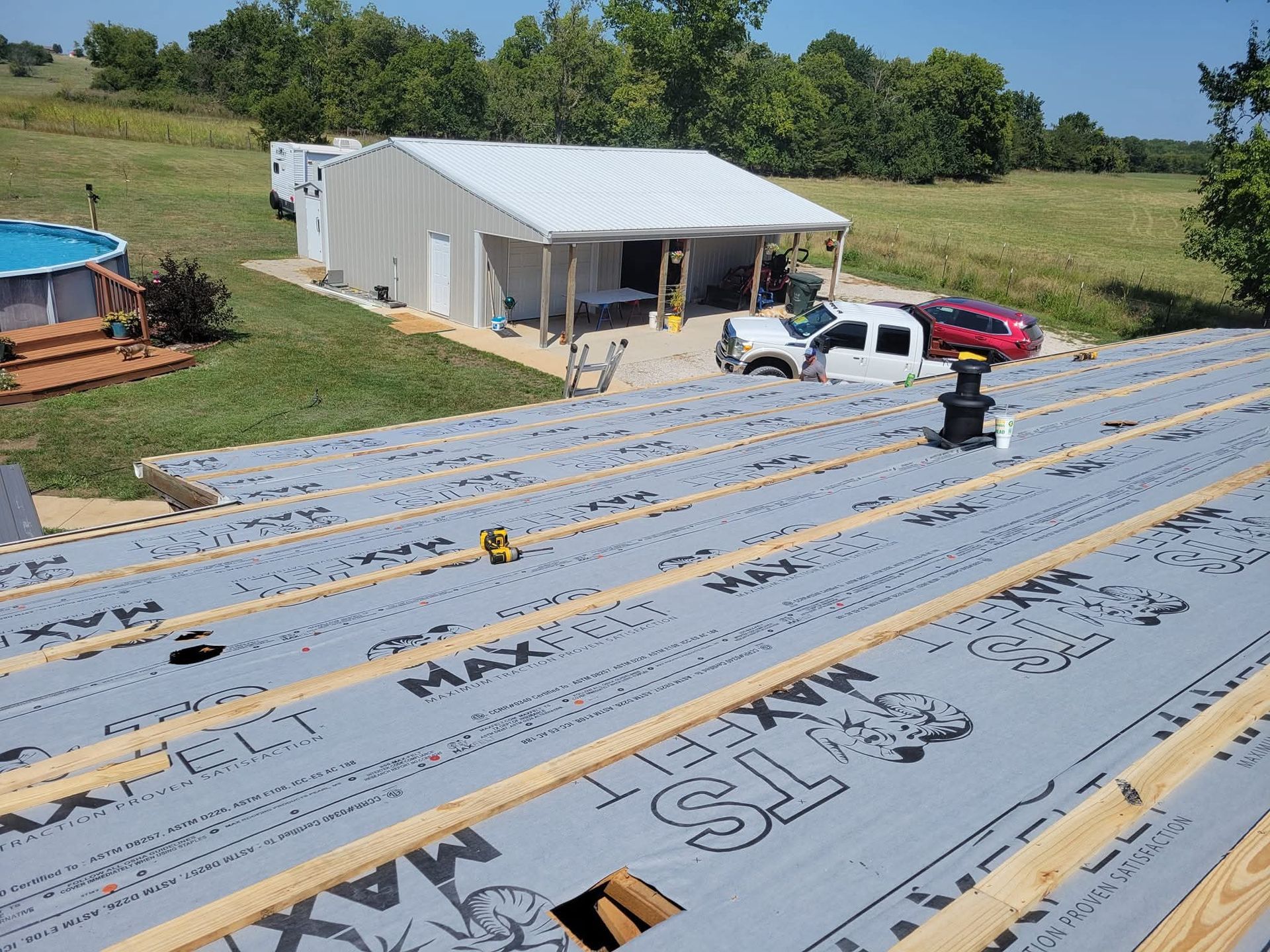Roof construction: Wooden beams and gray underlayment. White building and truck in background.
