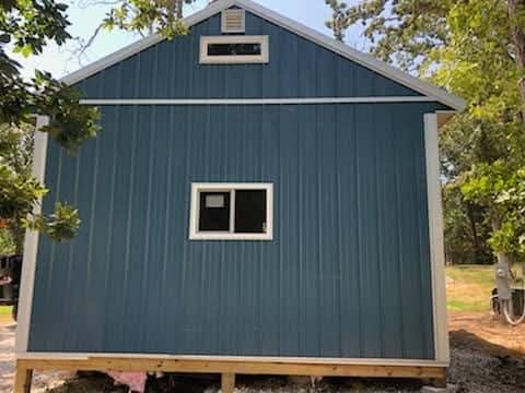 Blue metal-sided shed with white trim, a small window, and a rectangular vent.