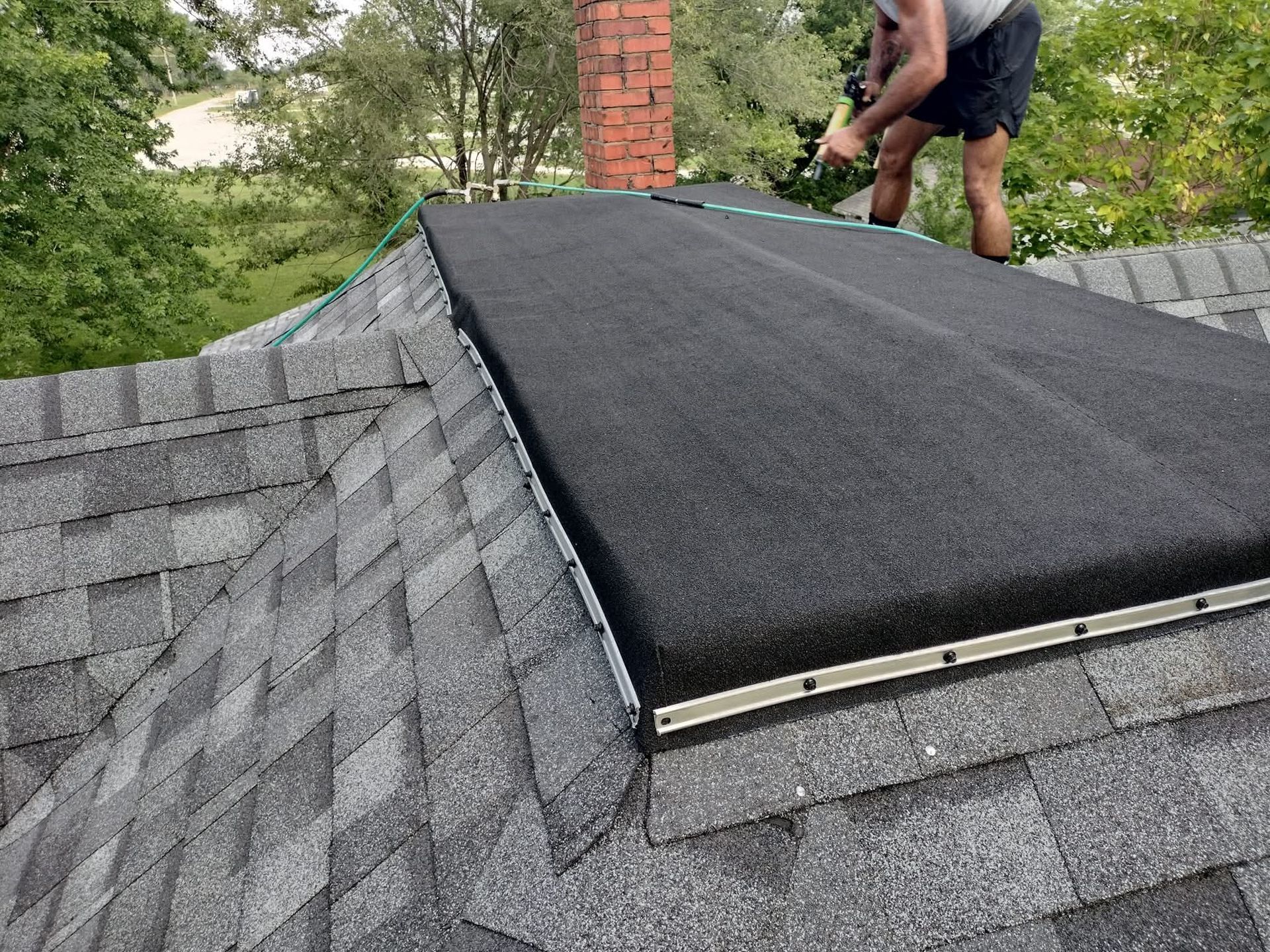 Man installing a flat, black roofing material on a roof near a chimney.