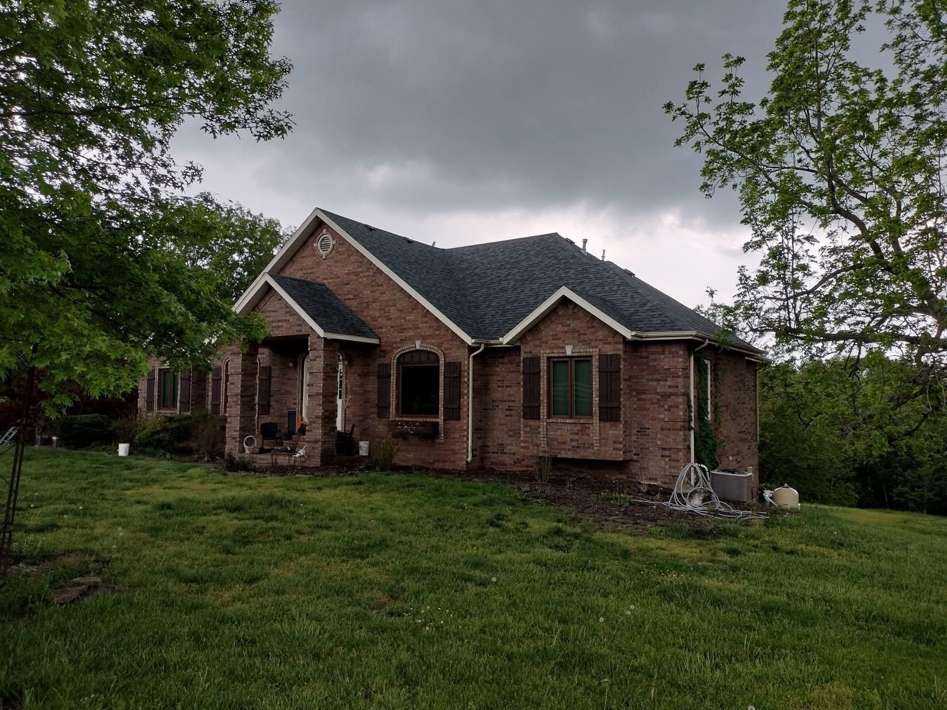Brick house with dark roof and brown shutters under a cloudy sky, surrounded by green trees and grass.