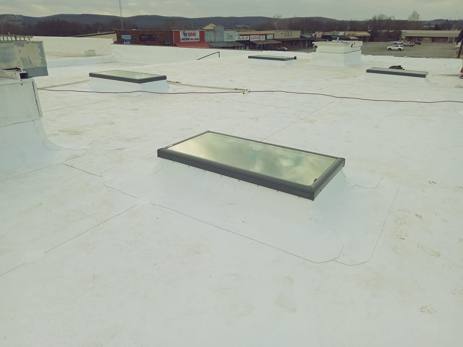 White commercial roof with rectangular skylights; overcast sky in the background.