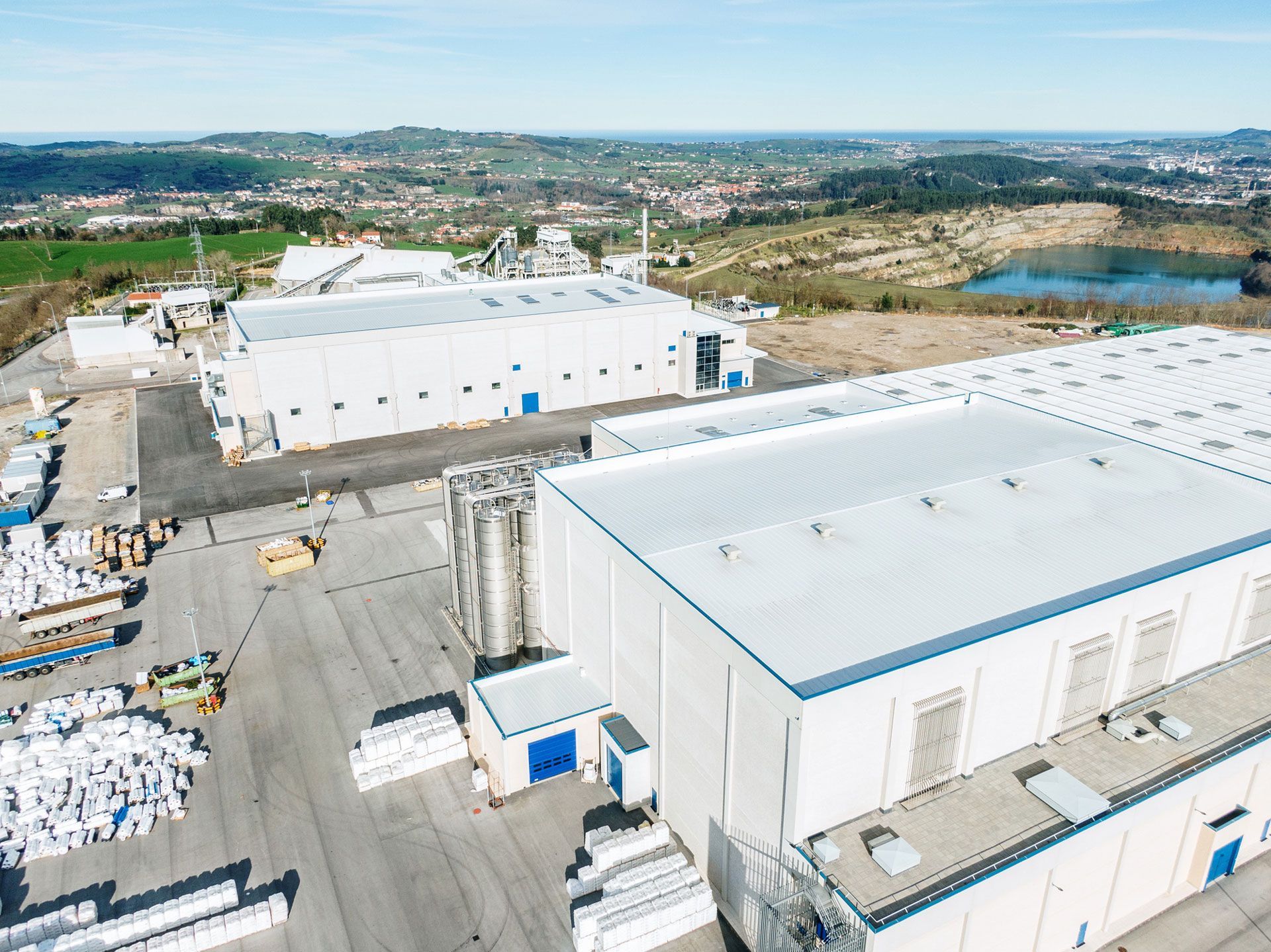Aerial view of an industrial complex with multiple white buildings, a paved lot, and a distant landscape with a lake.