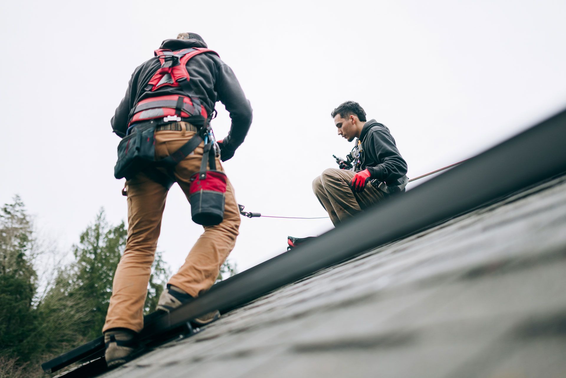 Two roofers on a rooftop; one is climbing, the other is sitting and looking down at something.
