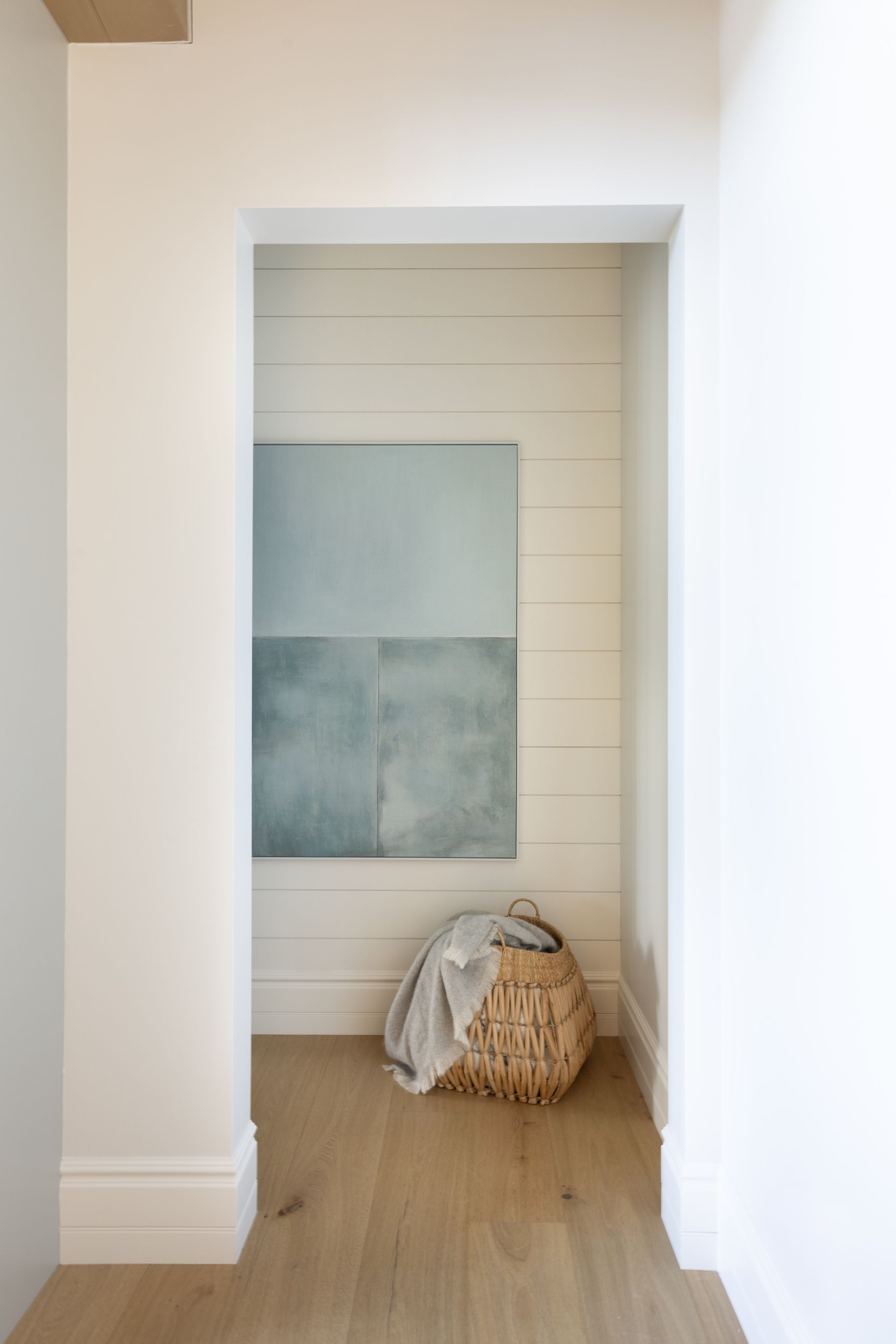 Minimalist hallway alcove with abstract wall art, wicker basket, and draped blanket on light wood floor