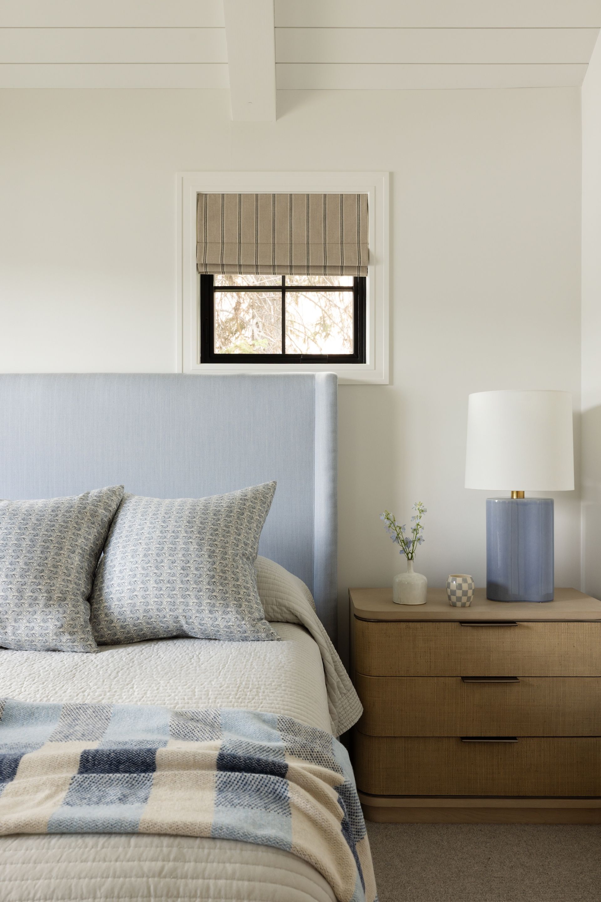 Bright bedroom with a blue upholstered bed, plaid blanket, wicker nightstand, and small window.