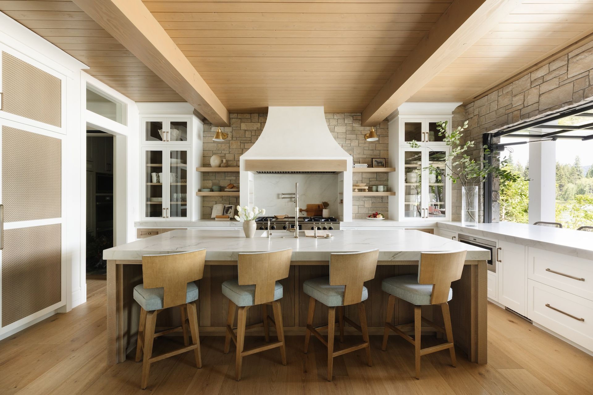 Bright modern kitchen with a large island, wooden stools, white cabinets, and stone accents