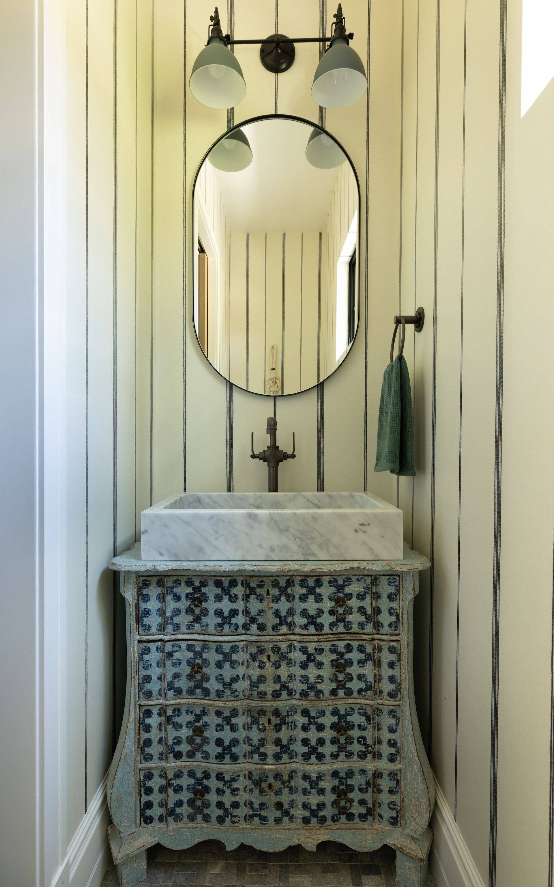 Narrow bathroom vanity with oval mirror, wall sconce, and tiled sink cabinet in a striped room