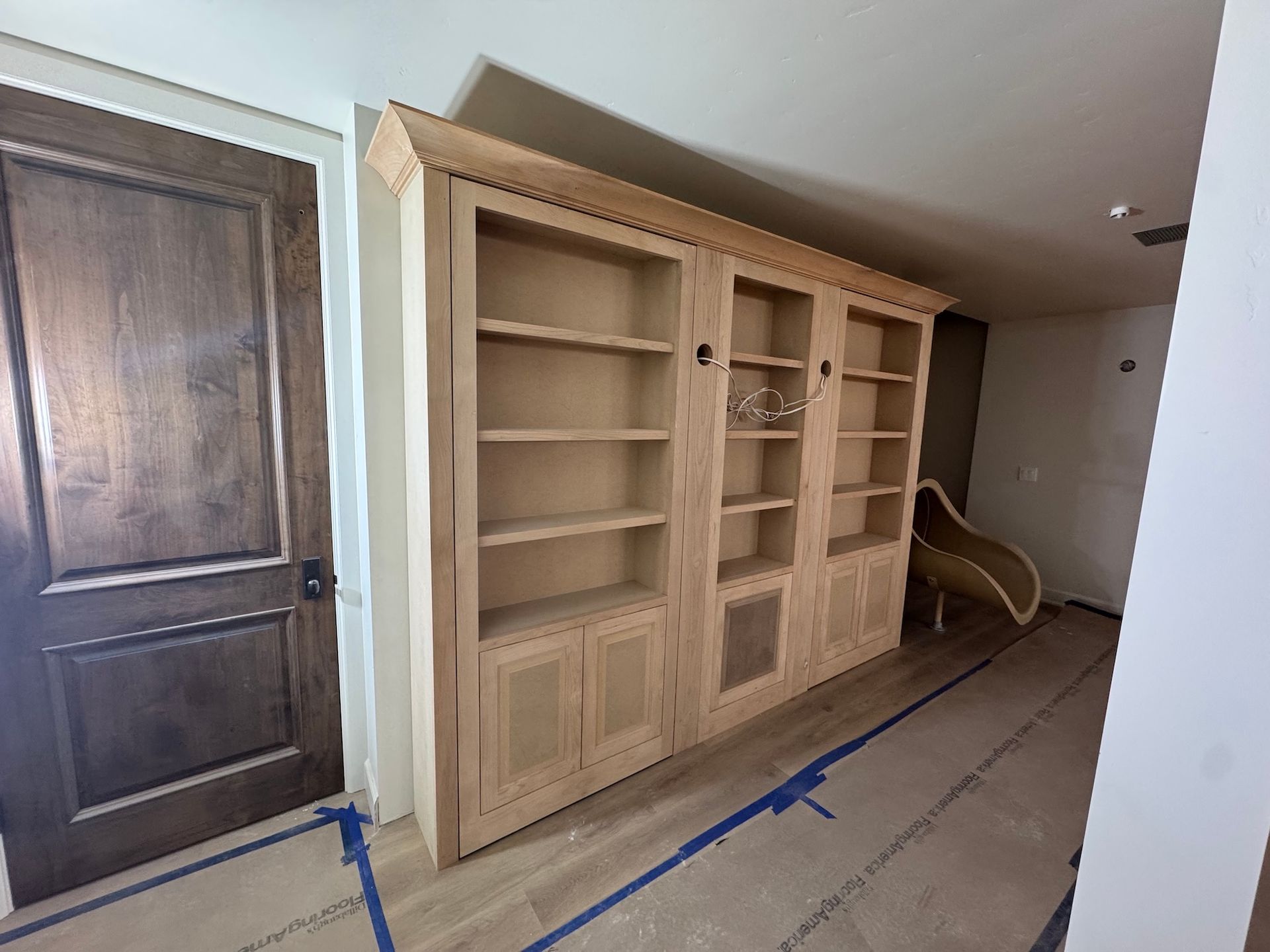 Built-in wooden cabinets and shelves in a hallway, with a door on the left and a chair in the back.