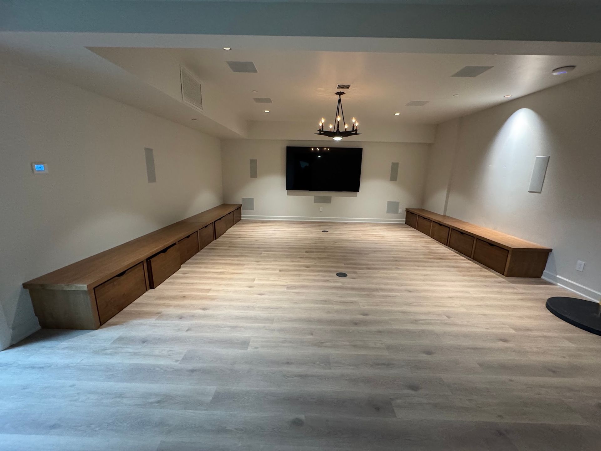 Empty conference room with long wooden benches, wood floor, wall-mounted TV, and chandelier lighting.