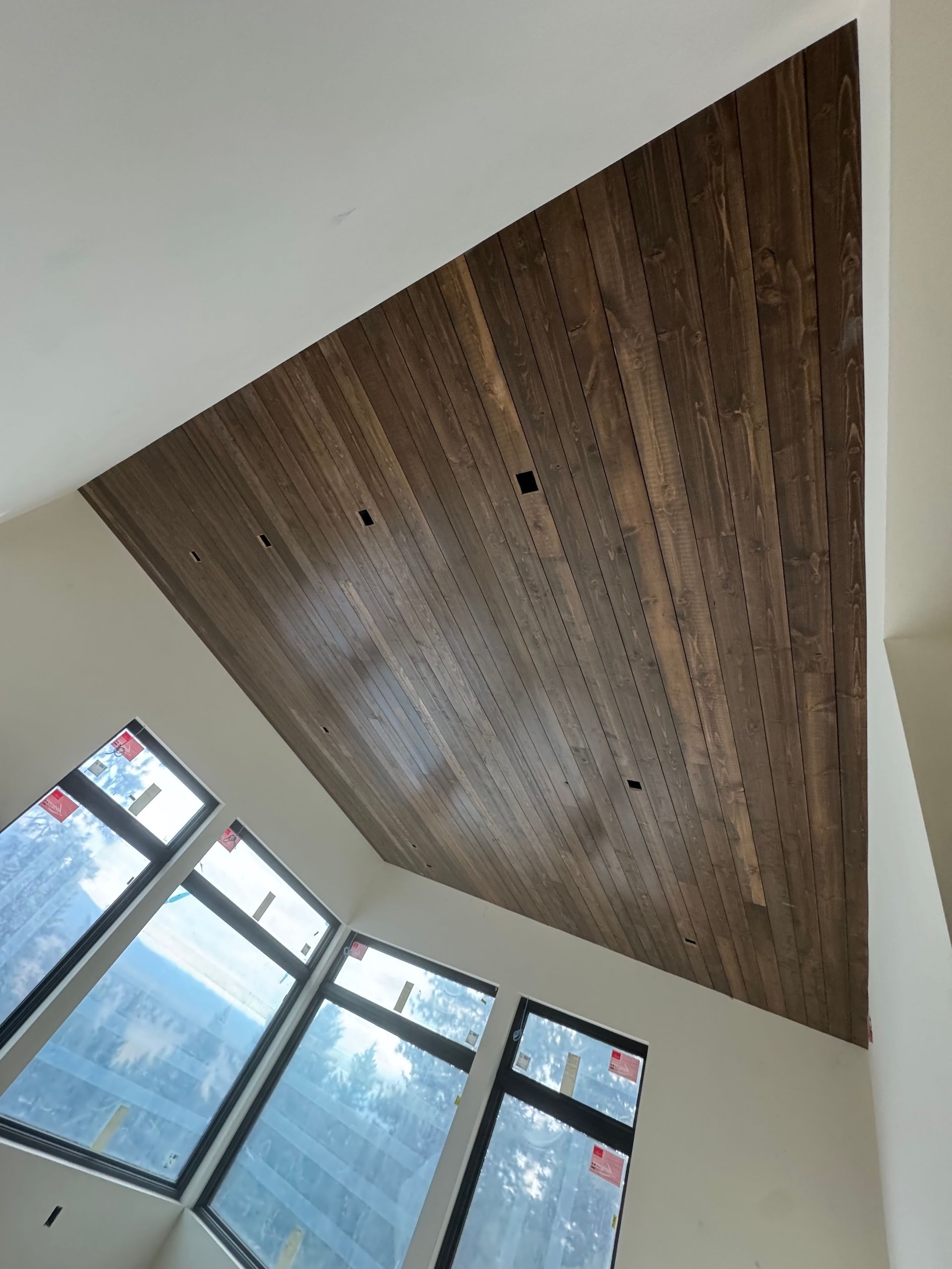 Angled view of a wood-paneled ceiling above tall windows in a bright interior.