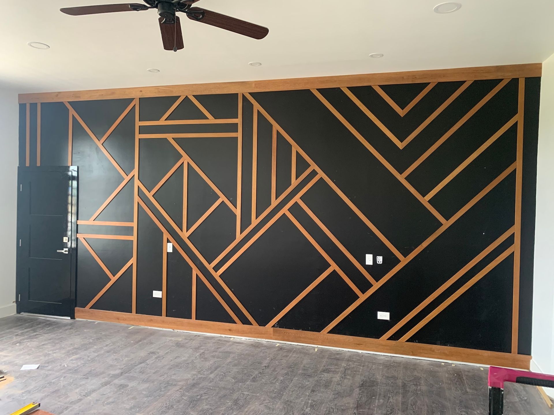 Black accent wall with tan wood geometric trim in an empty room with a ceiling fan and gray floor