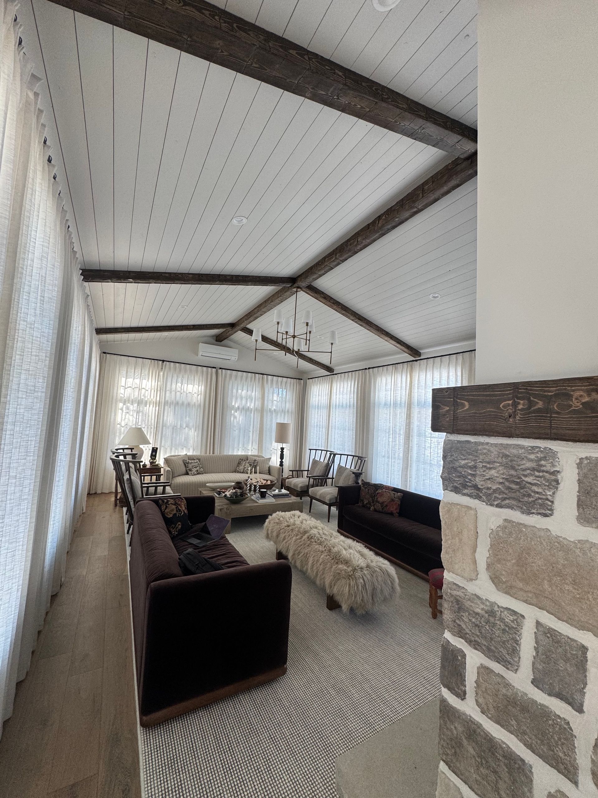 Bright sunroom living area with white curtains, dark sofas, stone fireplace, and a patterned rug.