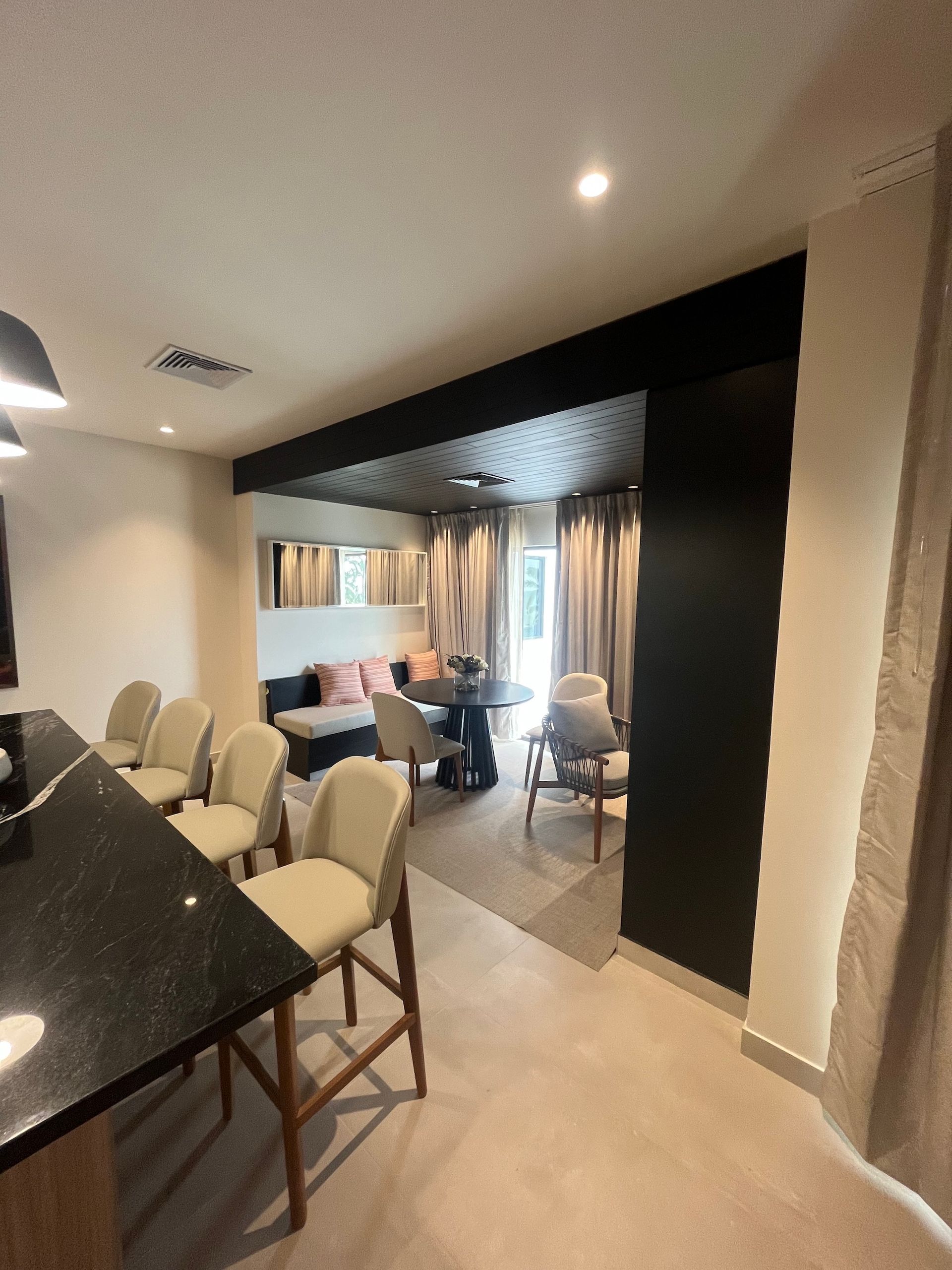 Modern dining room with a long black table, beige chairs, and a lounge area beyond a dark archway