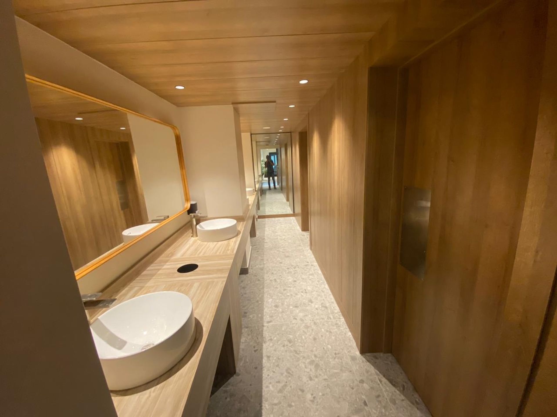 Long, modern wooden hallway with two white sinks, mirrors, and a view to a bright outdoor opening.