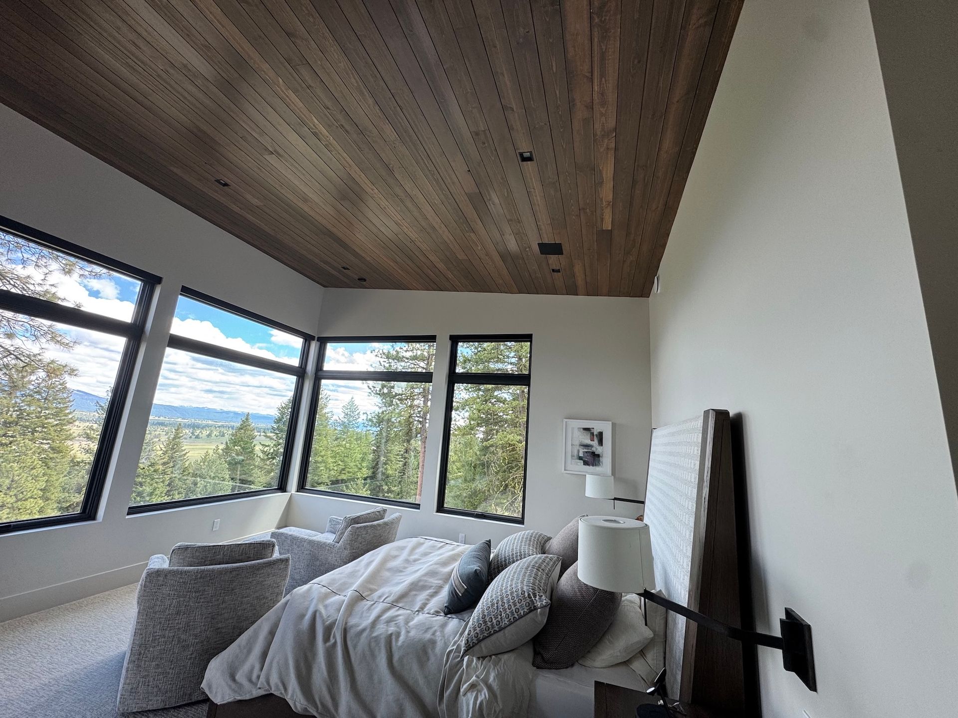 Modern bedroom with a wood ceiling, large windows, and a gray bed overlooking trees and mountains