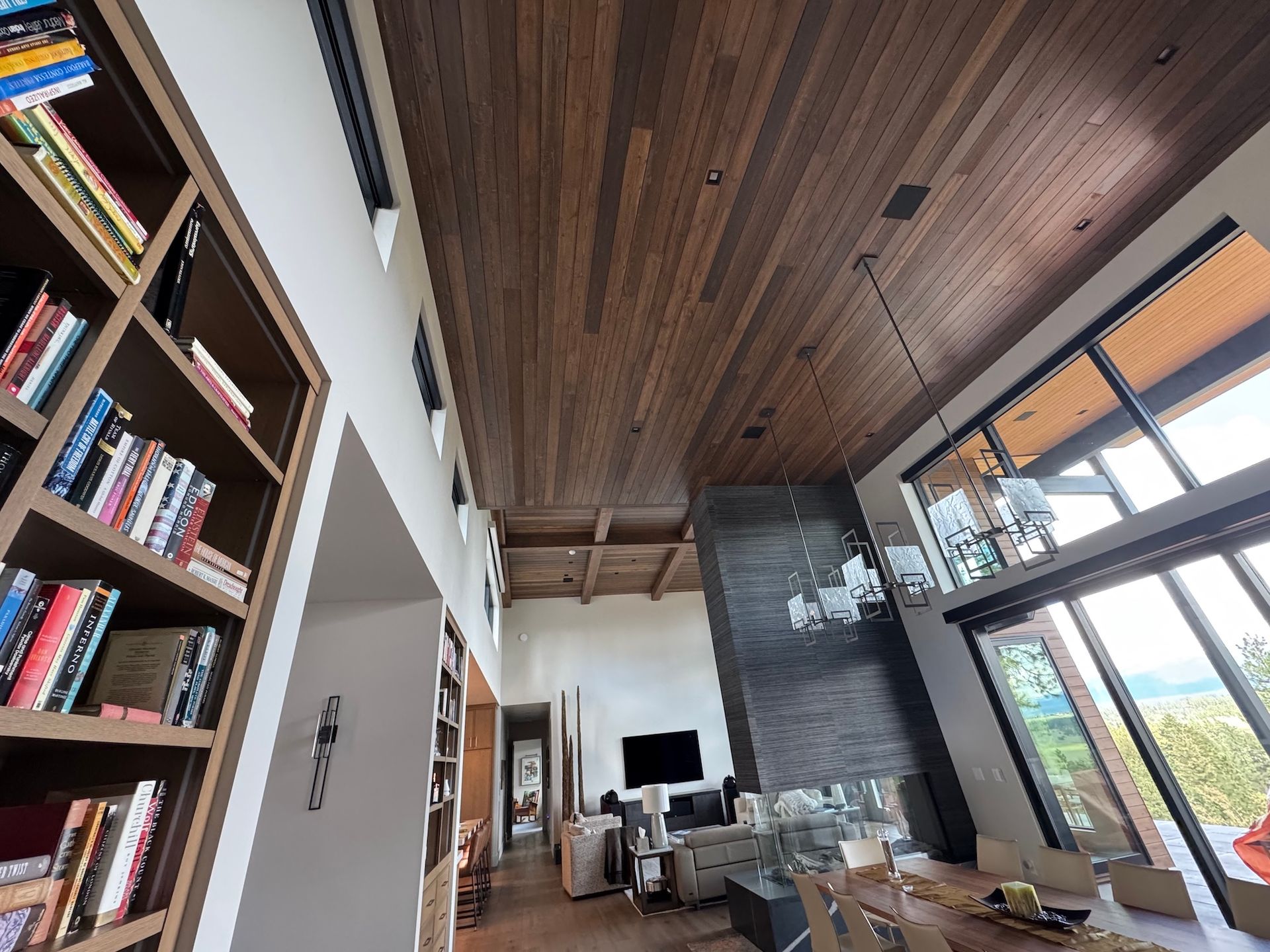 Modern living room with wood ceiling, tall bookshelves, fireplace, and large windows overlooking greenery