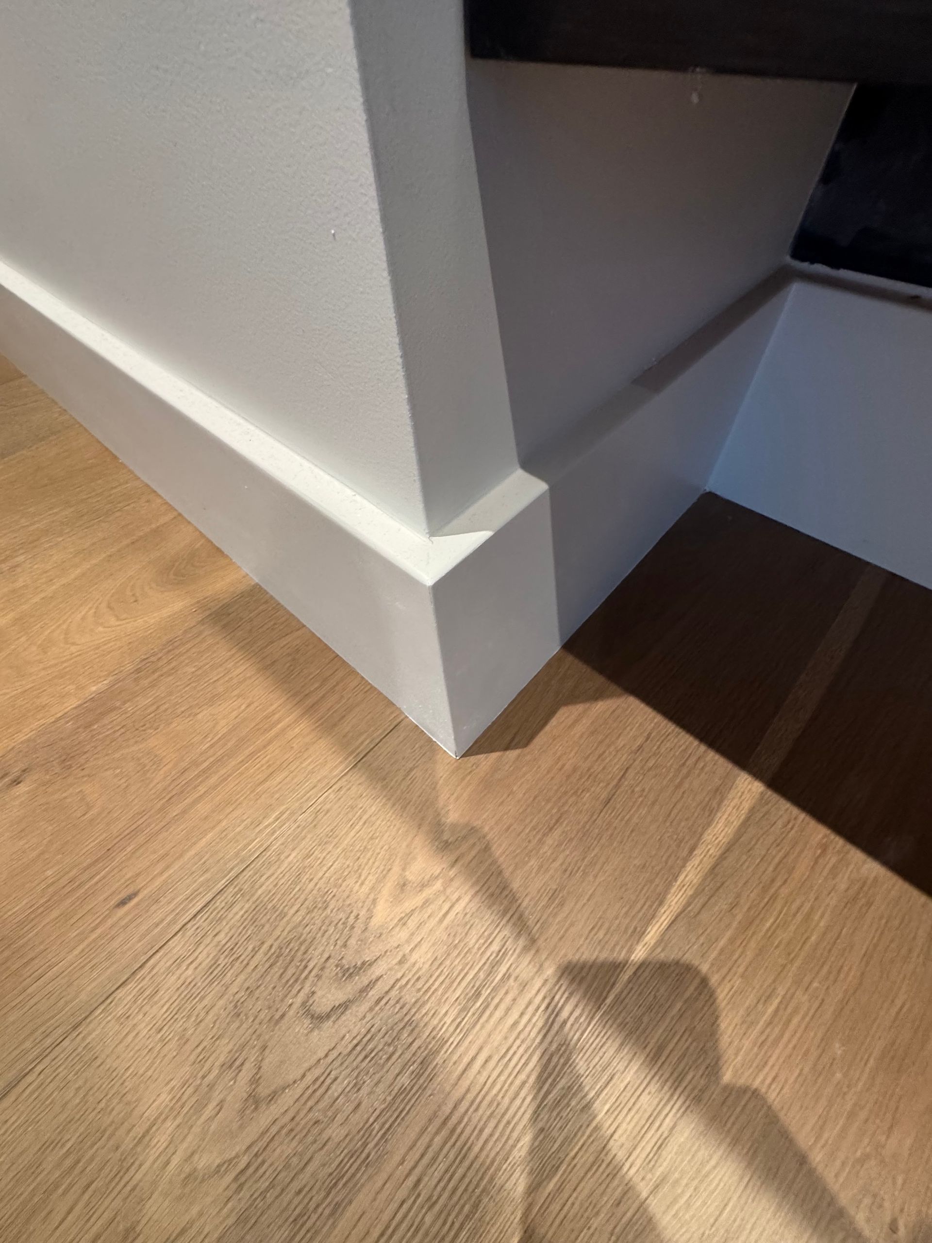 White corner trim on a wall beside a wood floor and stair opening, with sunlight casting shadows