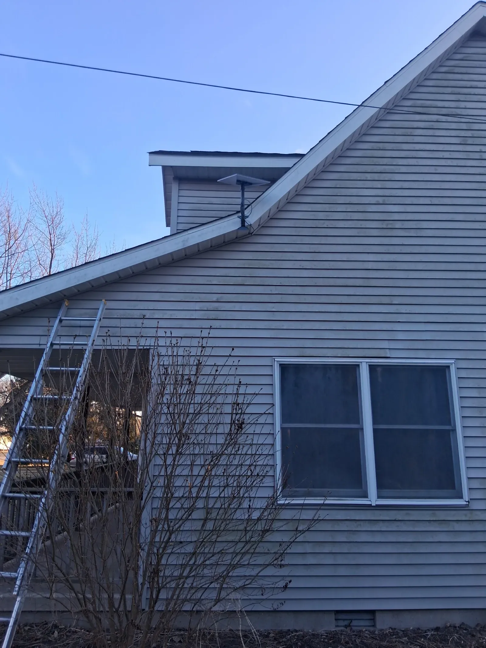 A ladder leans against the side of a grey house with peeling siding and an antenna attached near the roofline.