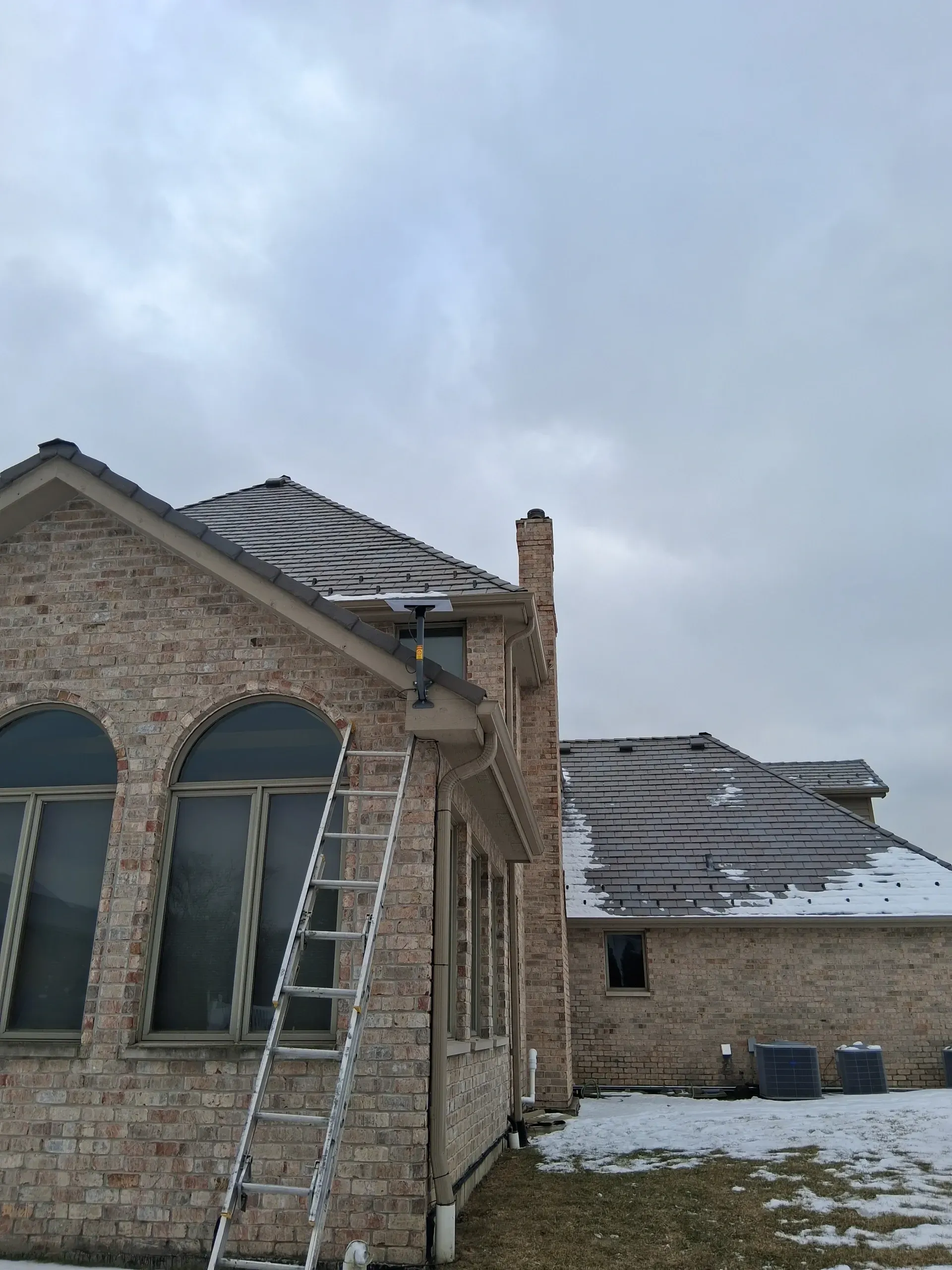 A ladder leans against a brick house with a TV antenna mounted near the roofline on a snowy day.
