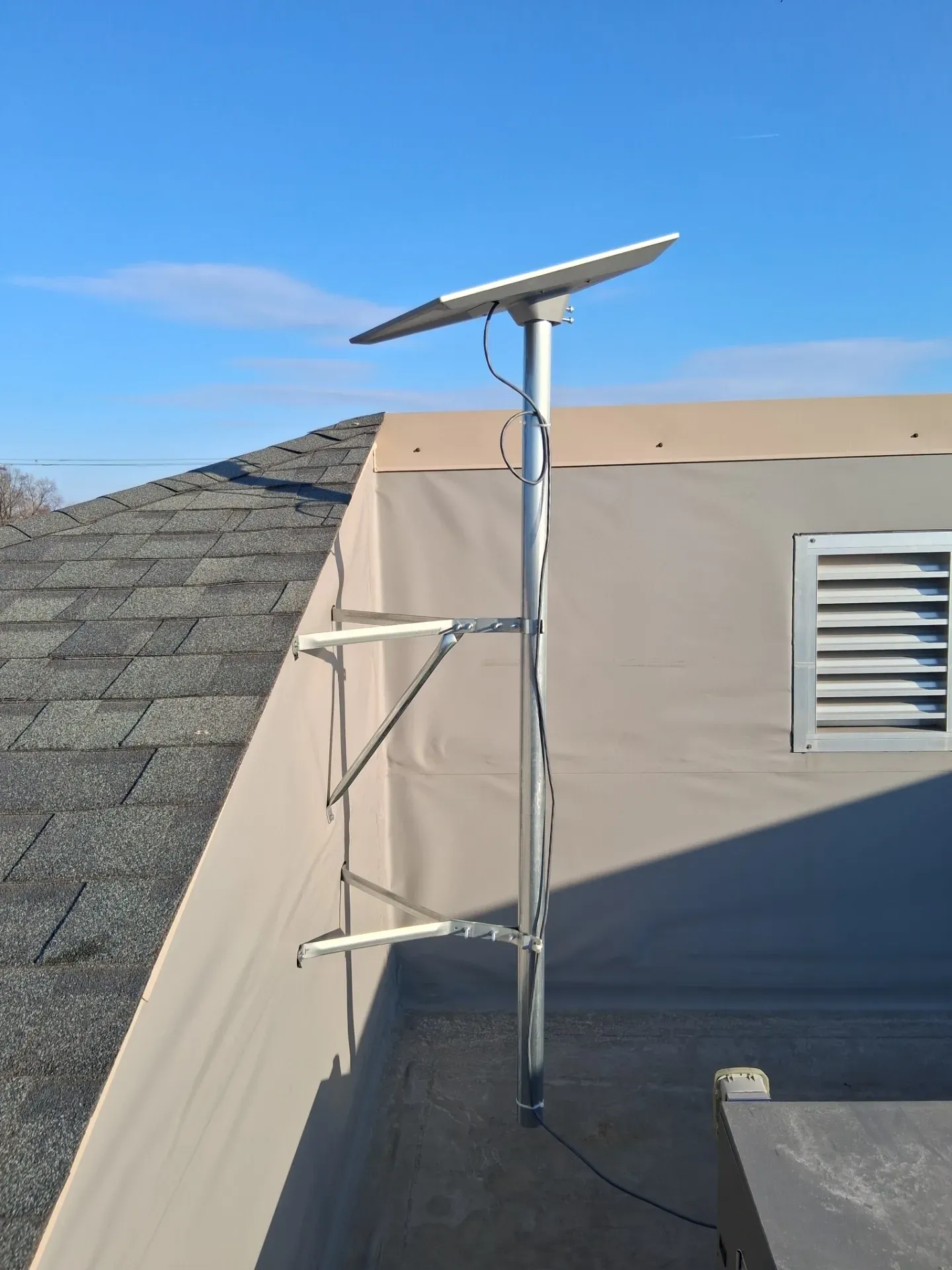 A Starlink satellite dish mounted on a vertical pole secured to the side of a building roof against a clear blue sky.