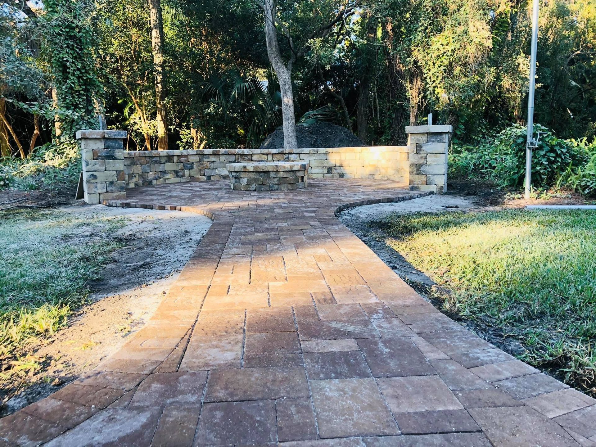 Brick walkway leading to a stone wall entrance, flanked by grass and trees in dappled sunlight