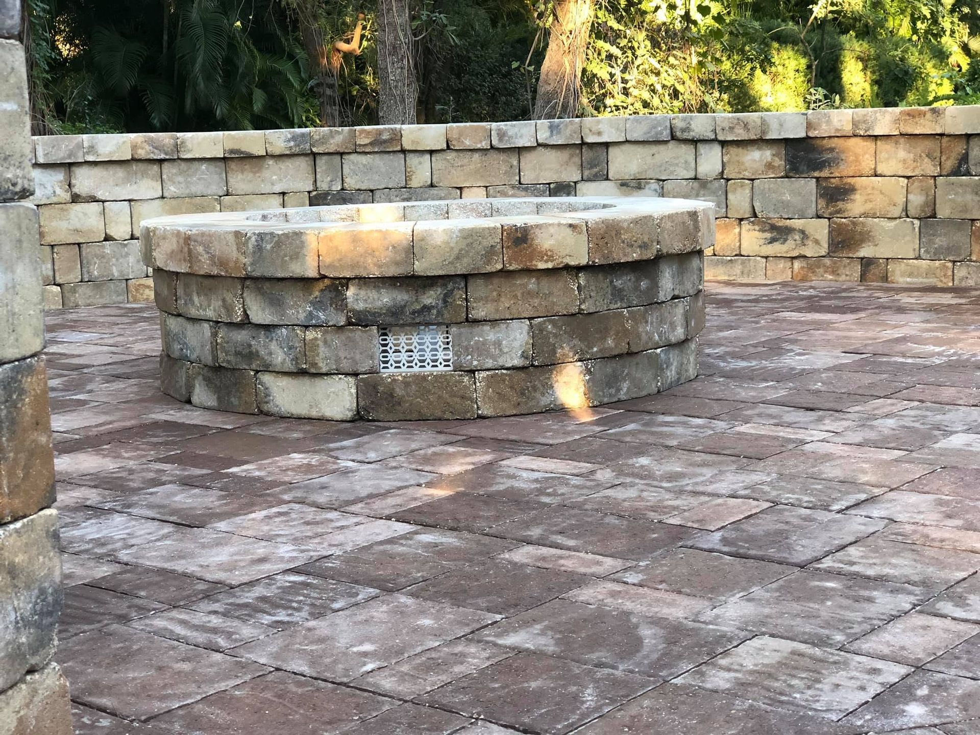 Stone fire pit in a paved courtyard, with a low stone wall and trees in the background.