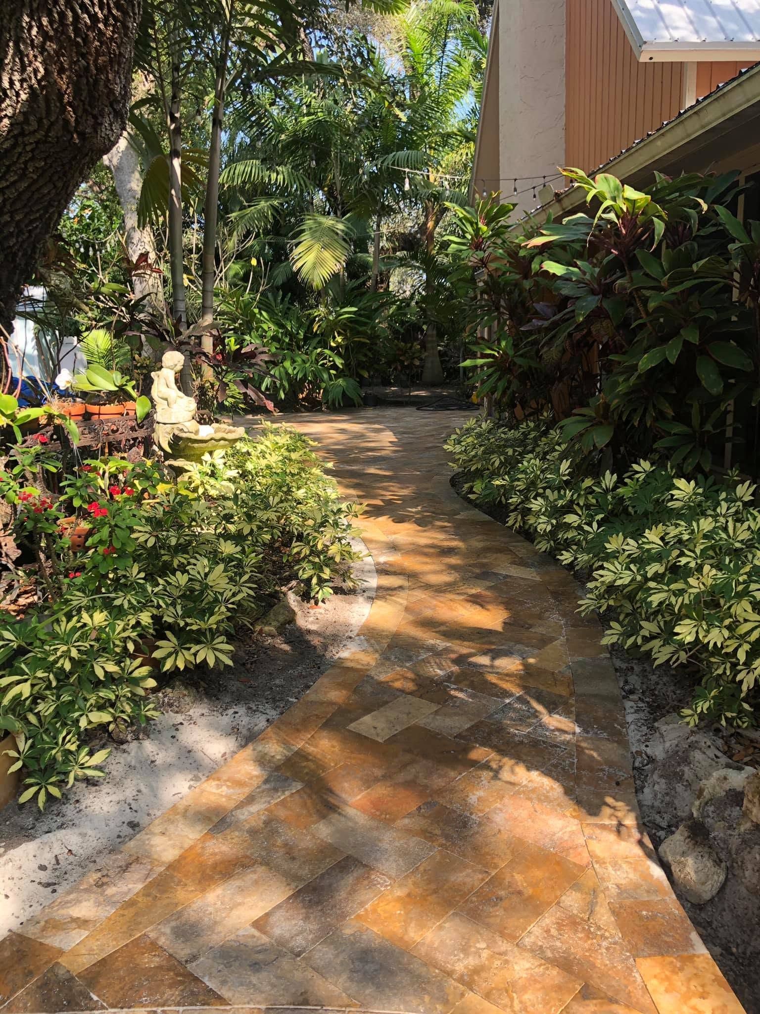 Brick garden path beside lush shrubs and a house, dappled in sunlight