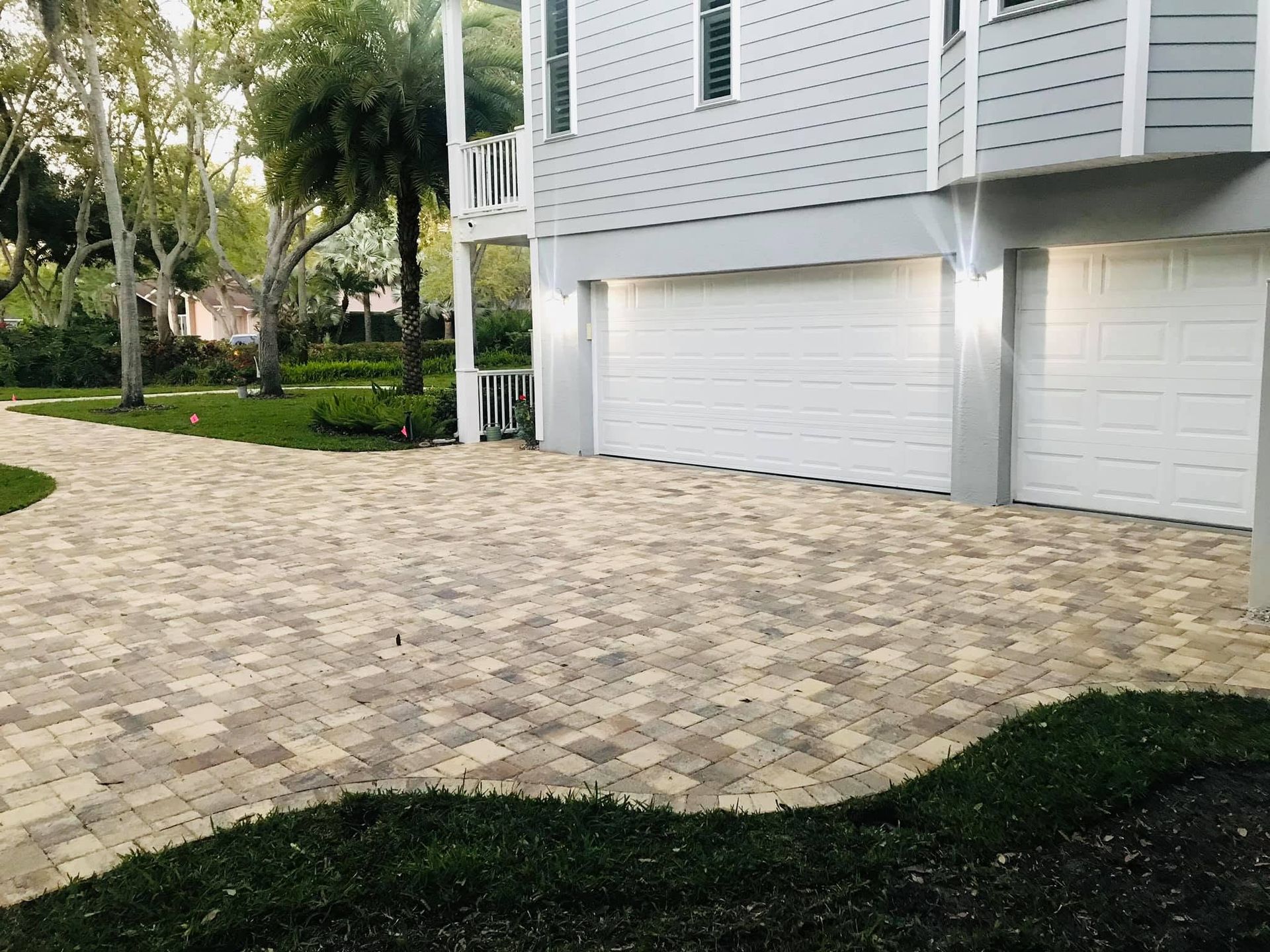 Driveway and walkway of light tan pavers beside a white house with garage doors and green lawn.