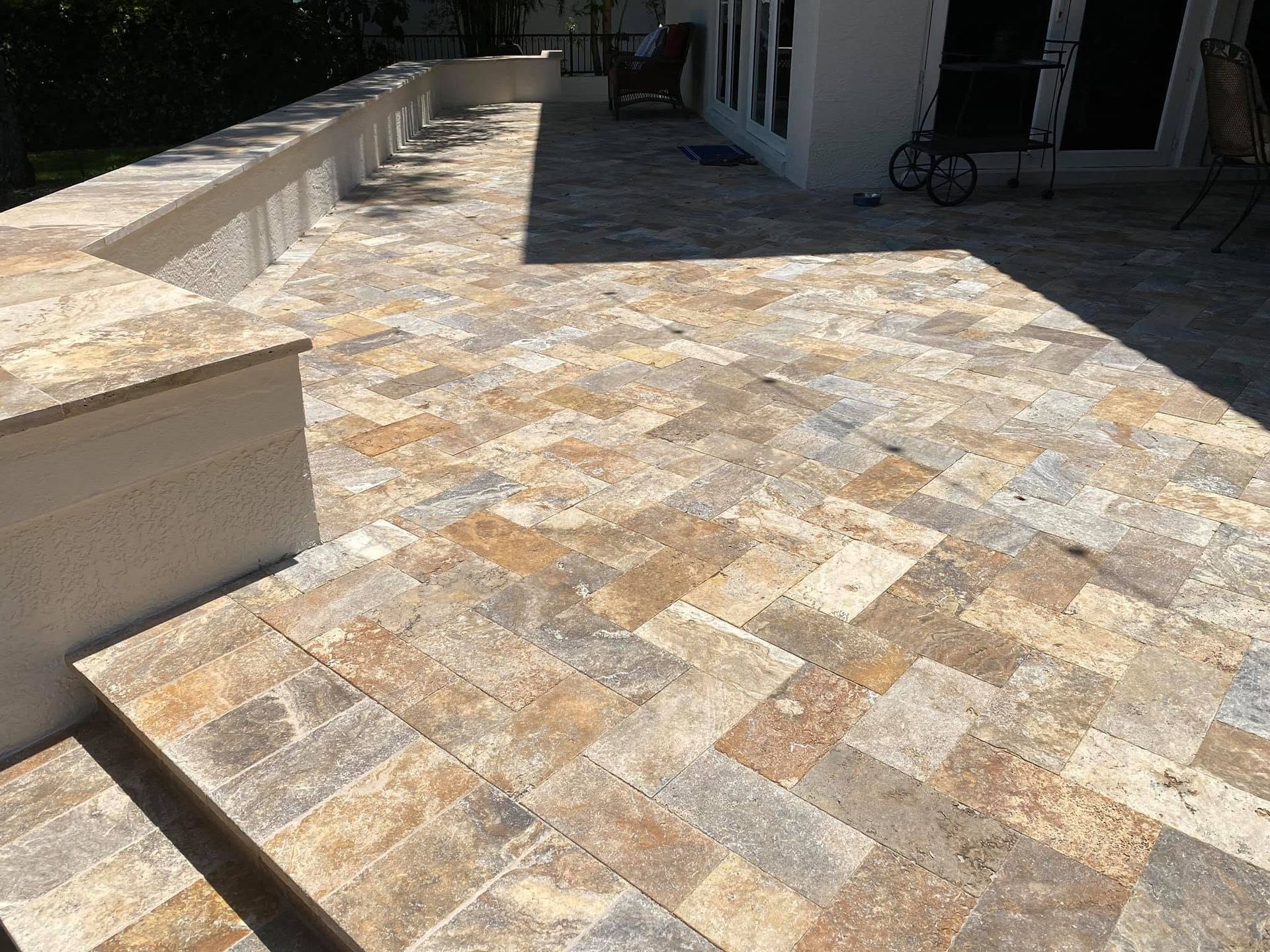 Sunlit stone patio with beige patterned pavers beside a white building and low wall