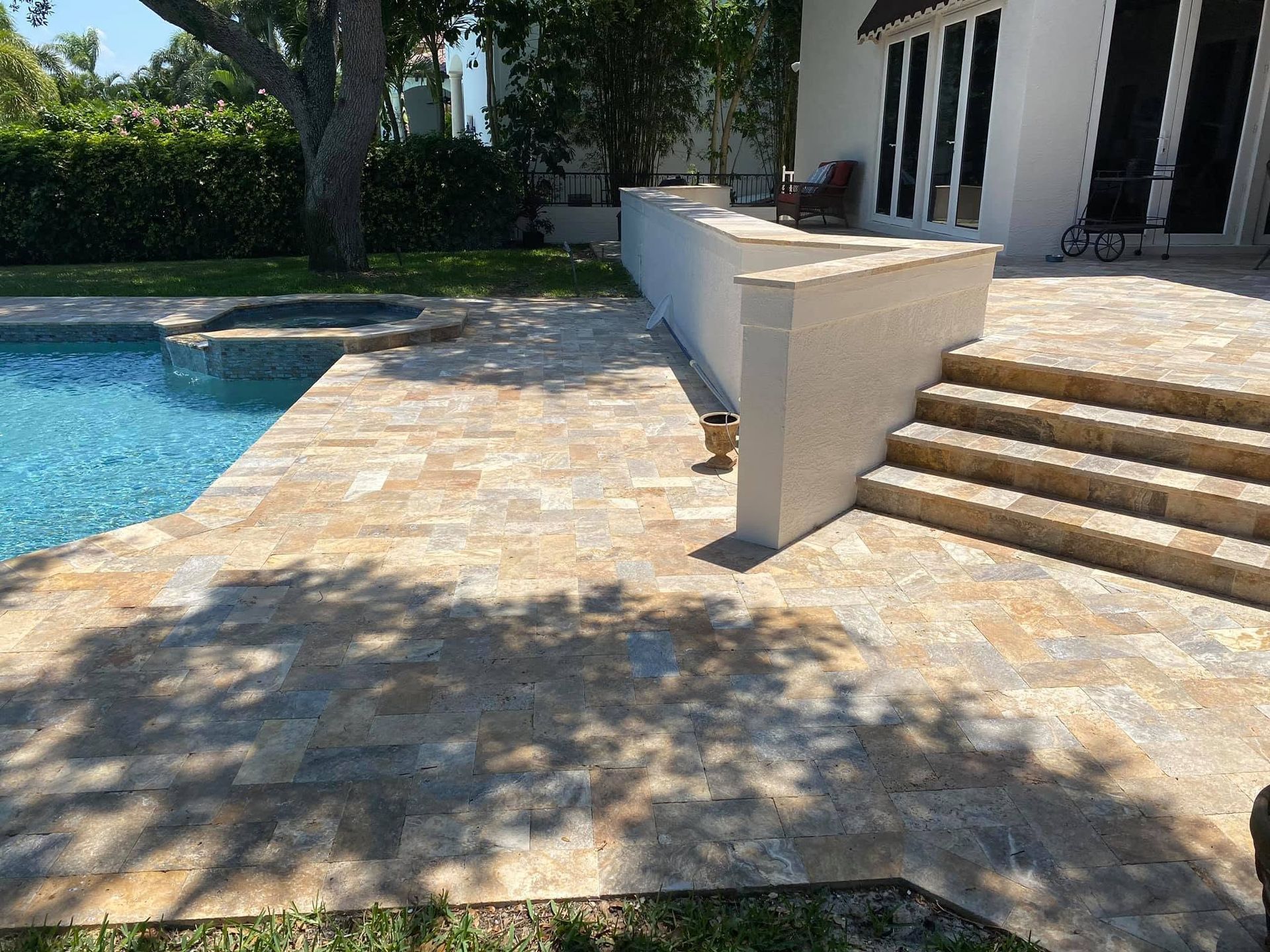 Stone patio beside a blue pool, with shaded trees and white house steps leading to a porch