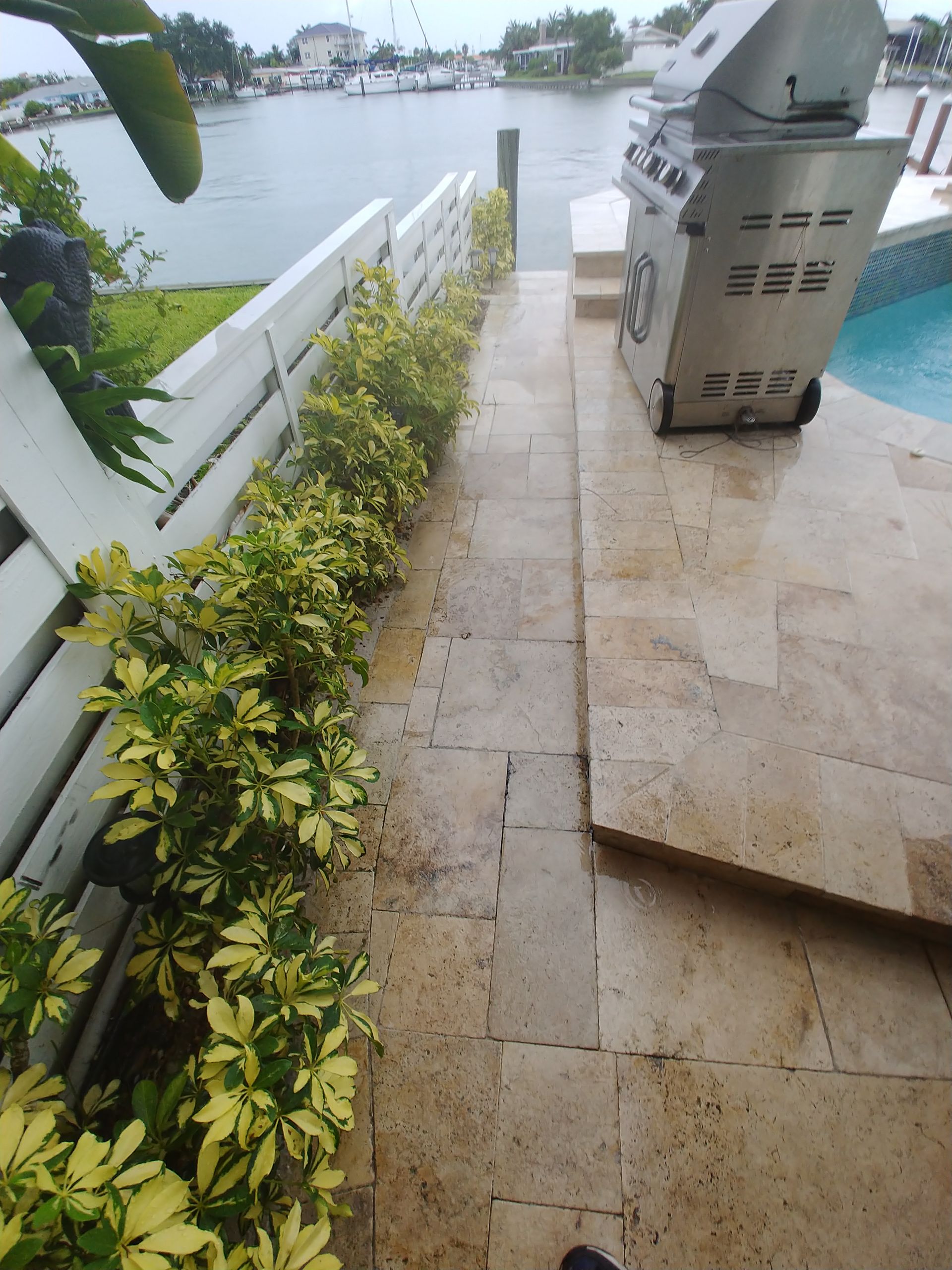 Narrow stone patio beside a waterfront, with a white railing, potted plants, and a grill overlooking the water.